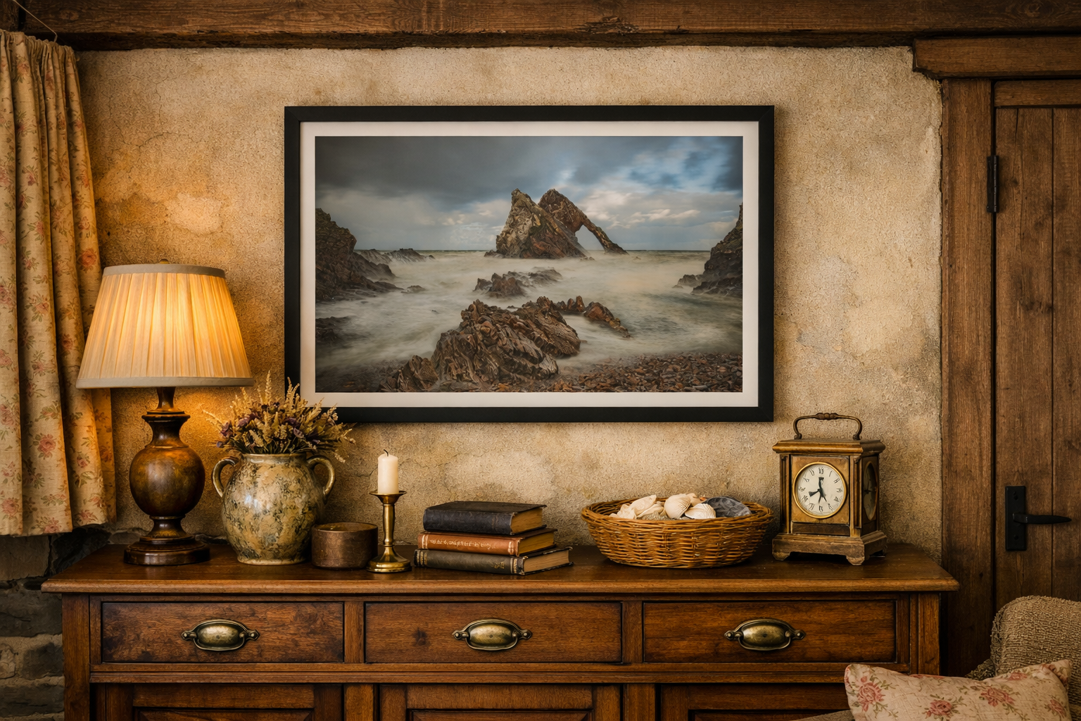 A rustic wooden sideboard decorated with a table lamp, a vase with flowers, books, a candle, a wicker basket with seashells, and a vintage clock. A framed coastal landscape photograph hangs above it. The room features textured beige walls, a floral curtain, and wooden accents.