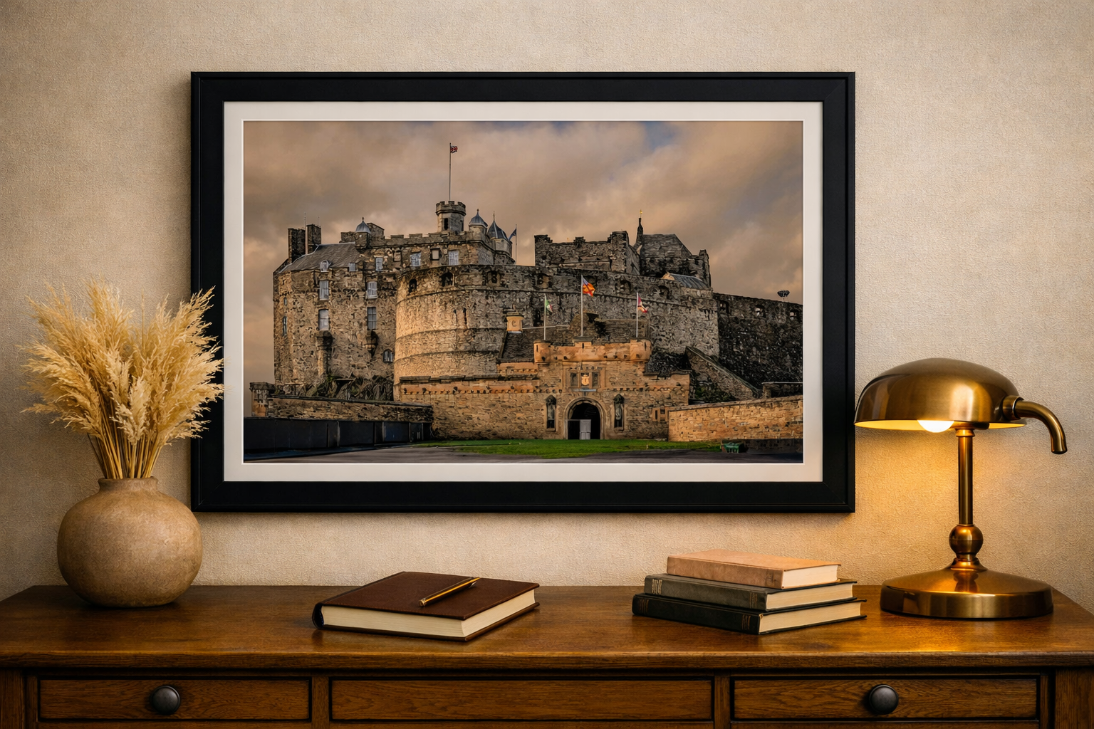 Framed photograph of a historic castle mounted on a beige wall above a wooden console table, decorated with a vase of dried pampas grass, a brass desk lamp, and a few stacked books.