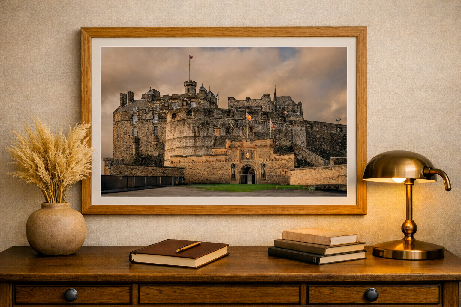 A framed photograph of a medieval castle hanging on a beige wall, with a wooden desk below it. The desk has a brown book with a pen on top, a gold desk lamp, and a stack of four books. A beige vase with dried wheat stalks is on the left side of the desk.