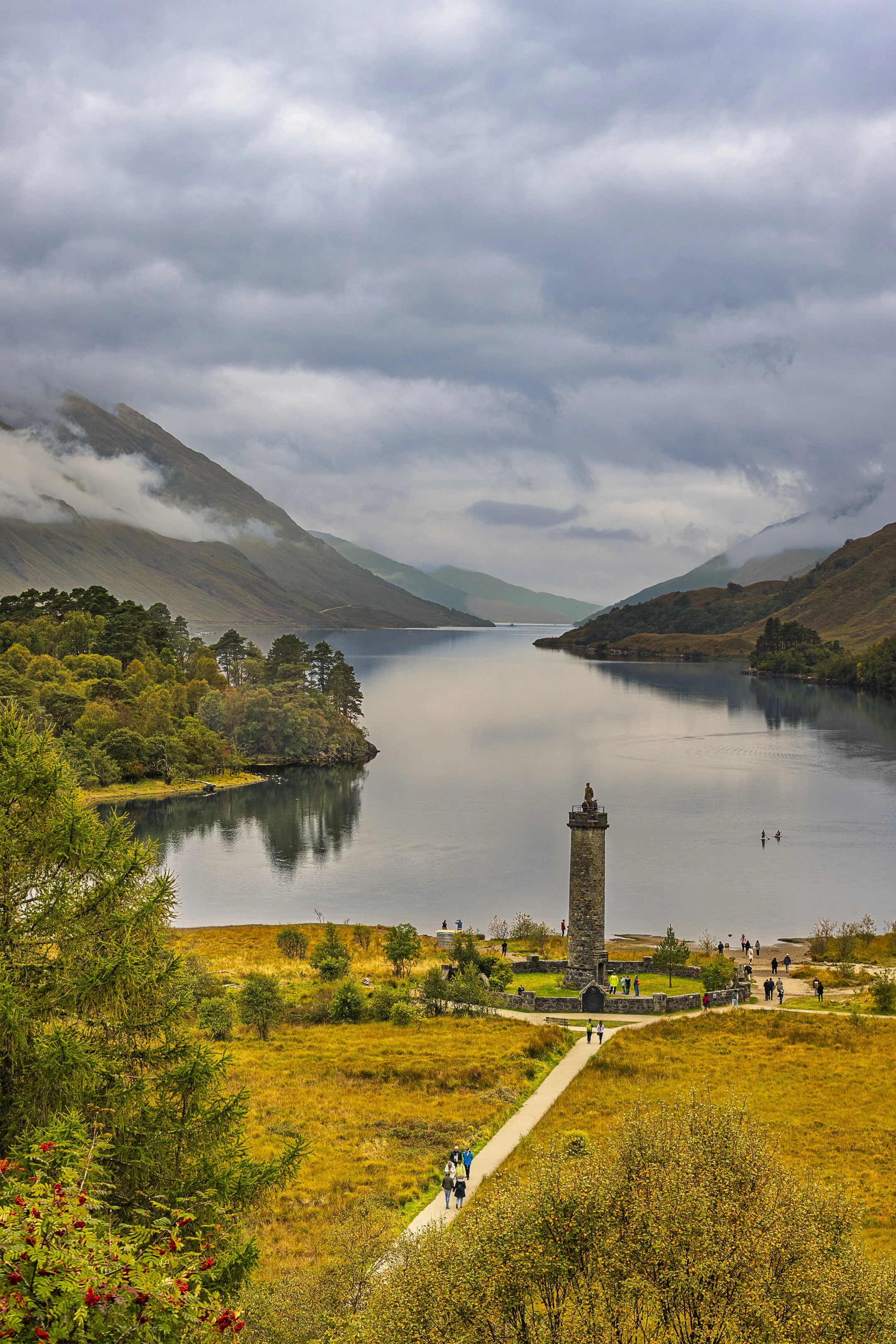 A scenic view of a lake surrounded by mountains with clouds overhead. A pathway leads to a stone tower with people around it, and a few boats are on the lake.