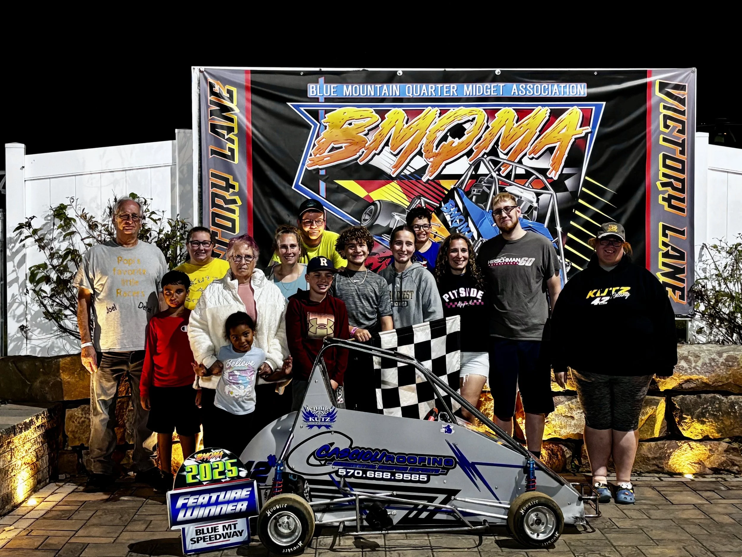 A group of people, including children and adults, gathered outdoors at night in front of a banner for Blue Mountain Quarter Midget Association at Blue MT Speedway. A small race car with sponsor logos and a checkered flag pattern on the front is in the foreground.