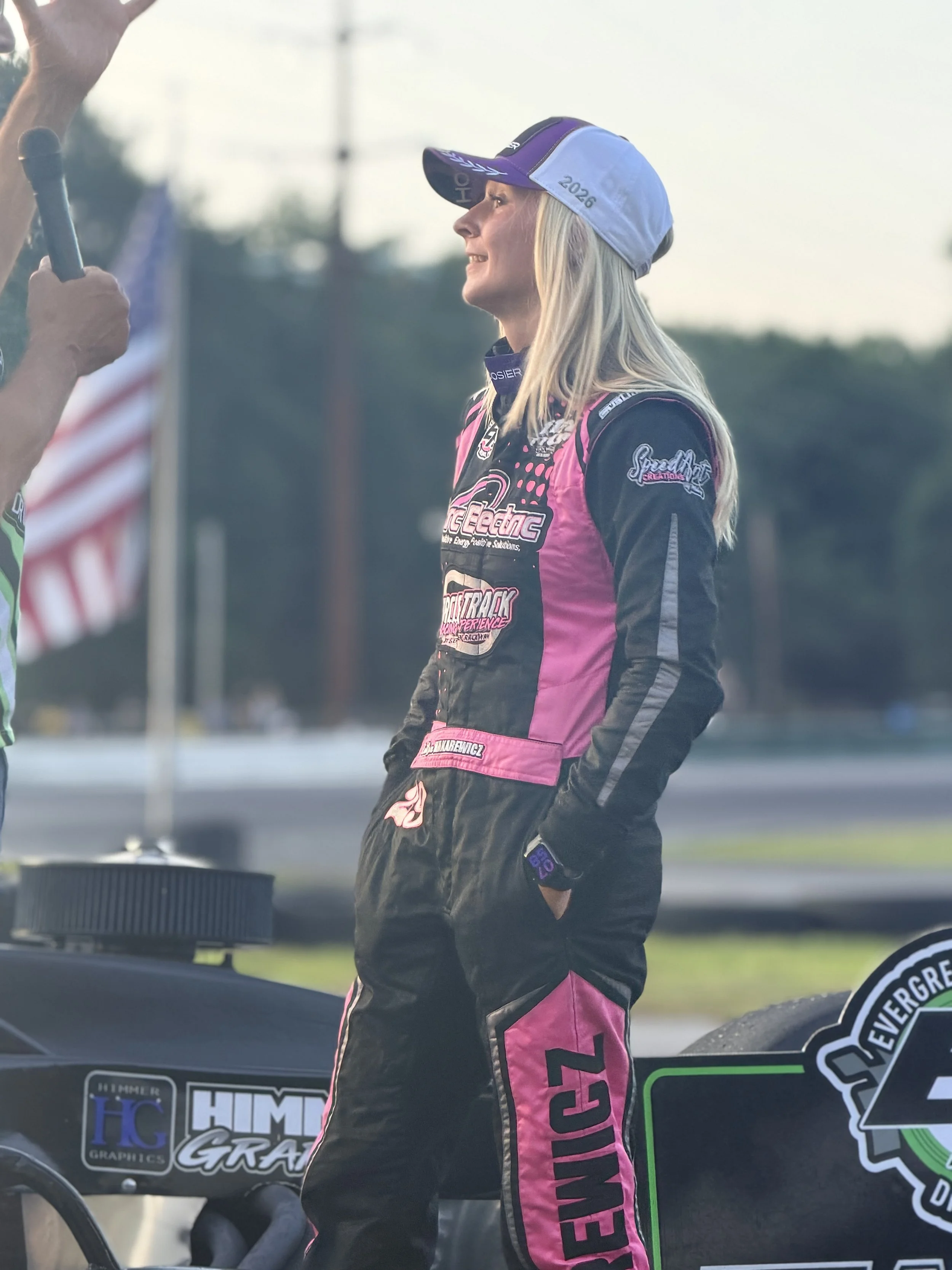 A female race car driver wearing a racing suit and a cap with the year 2026 on it, standing next to her race car and speaking to someone.
