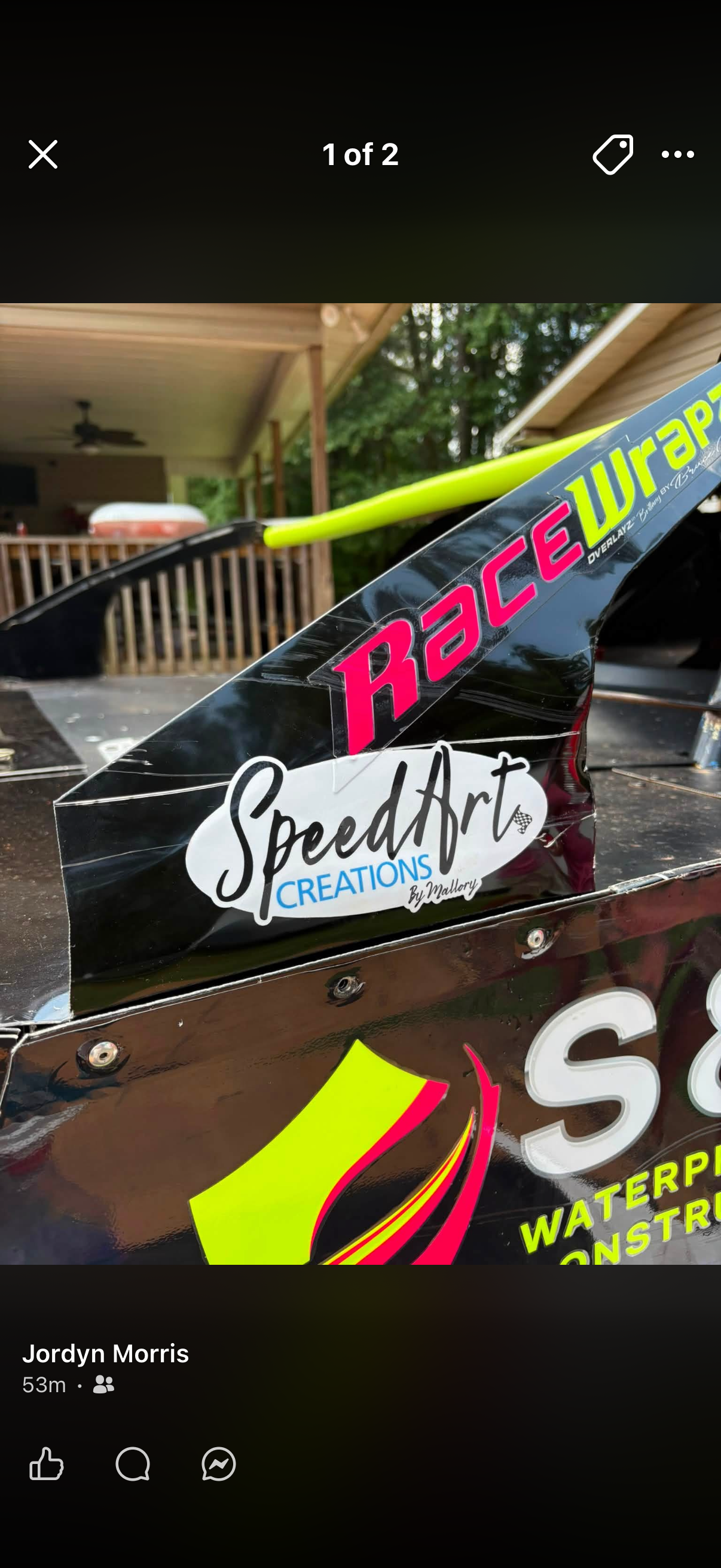 Close-up of a black racing car wing with colorful decals showing the words 'RACE Wraph,' 'SpeedArt Creations by Mallory,' and parts of a watercraft construction project in the background.