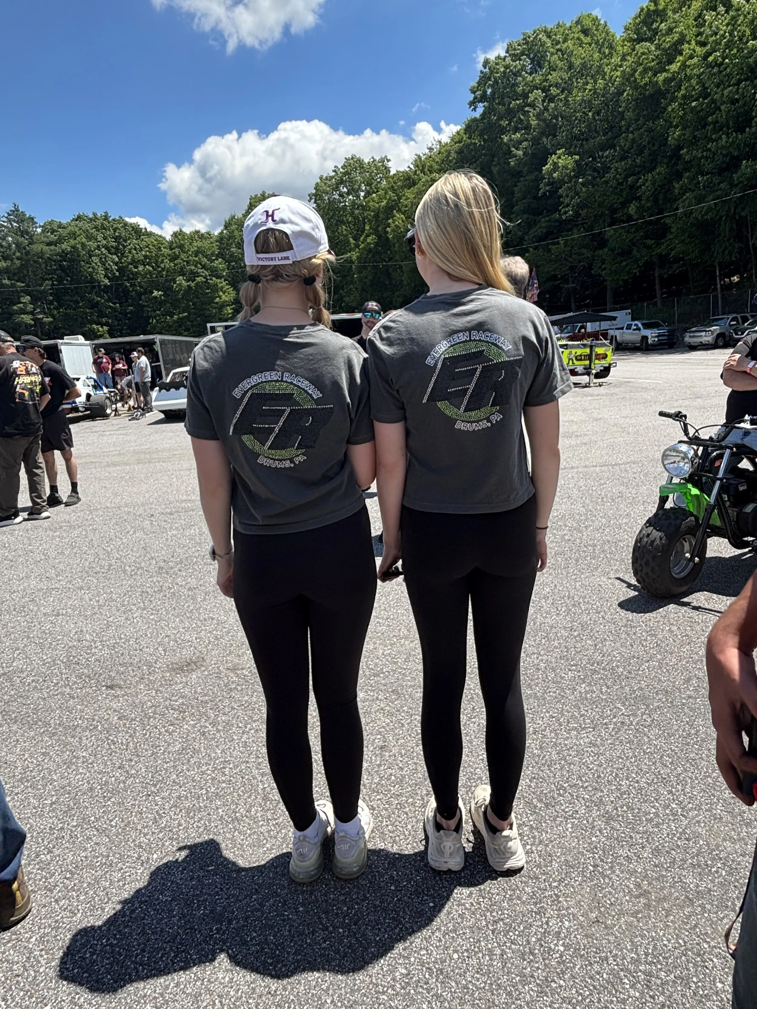 Two women are standing outside, facing away from the camera. They are wearing matching black leggings, white sneakers, and gray T-shirts with a logo that says "Evergreen Raceway" and "Drums, PA." One woman is wearing a white baseball cap, and the other has long blonde hair. There are other people, vehicles, and trees in the background on a sunny day.