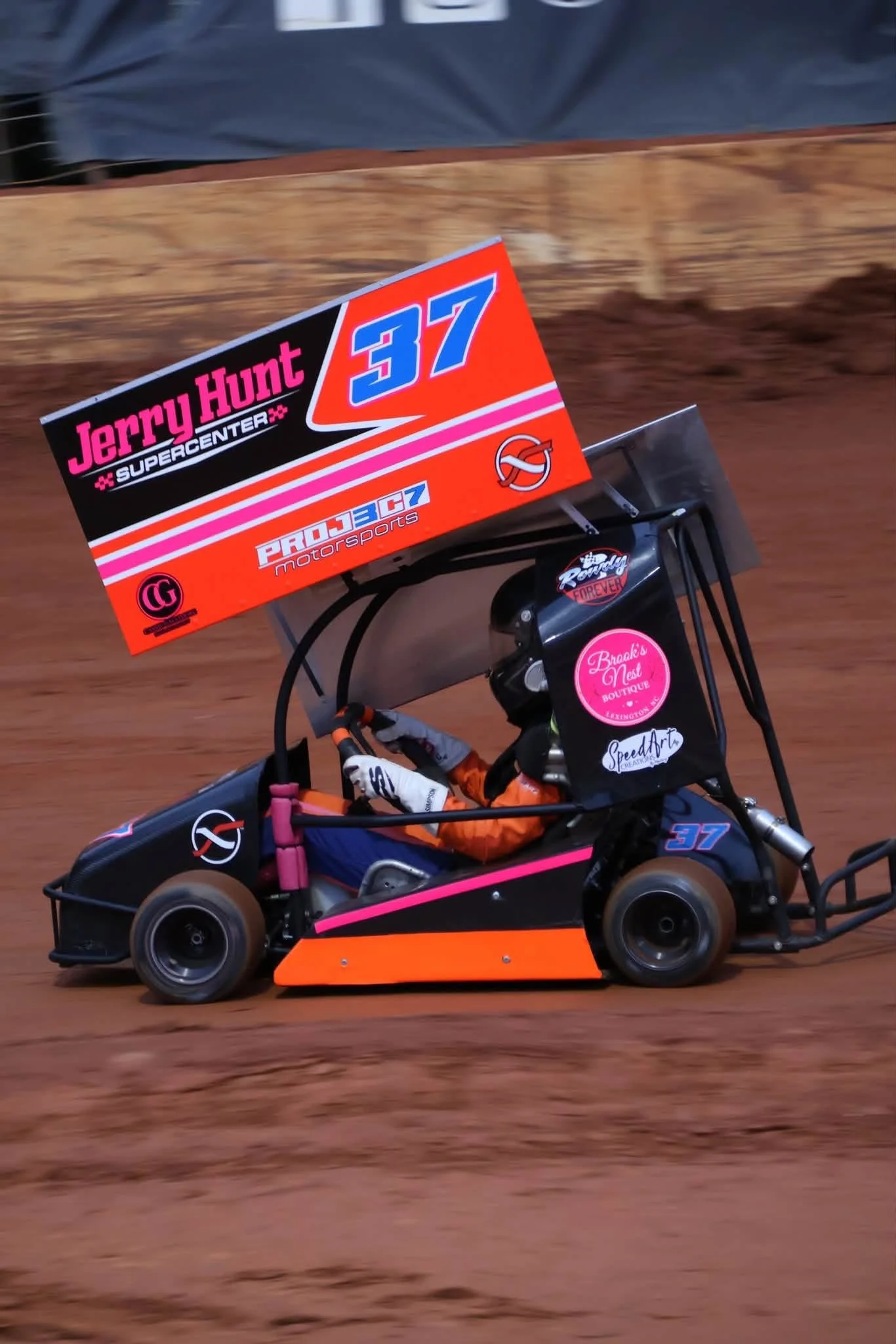 A small race car with a black, pink, orange, and blue color scheme speeds around a dirt track. The car has a large rear wing with sponsor logos, including 'Jerry Hunt Supercenter' and 'Pro J3 Sports.' The driver is wearing a black helmet and racing suit.