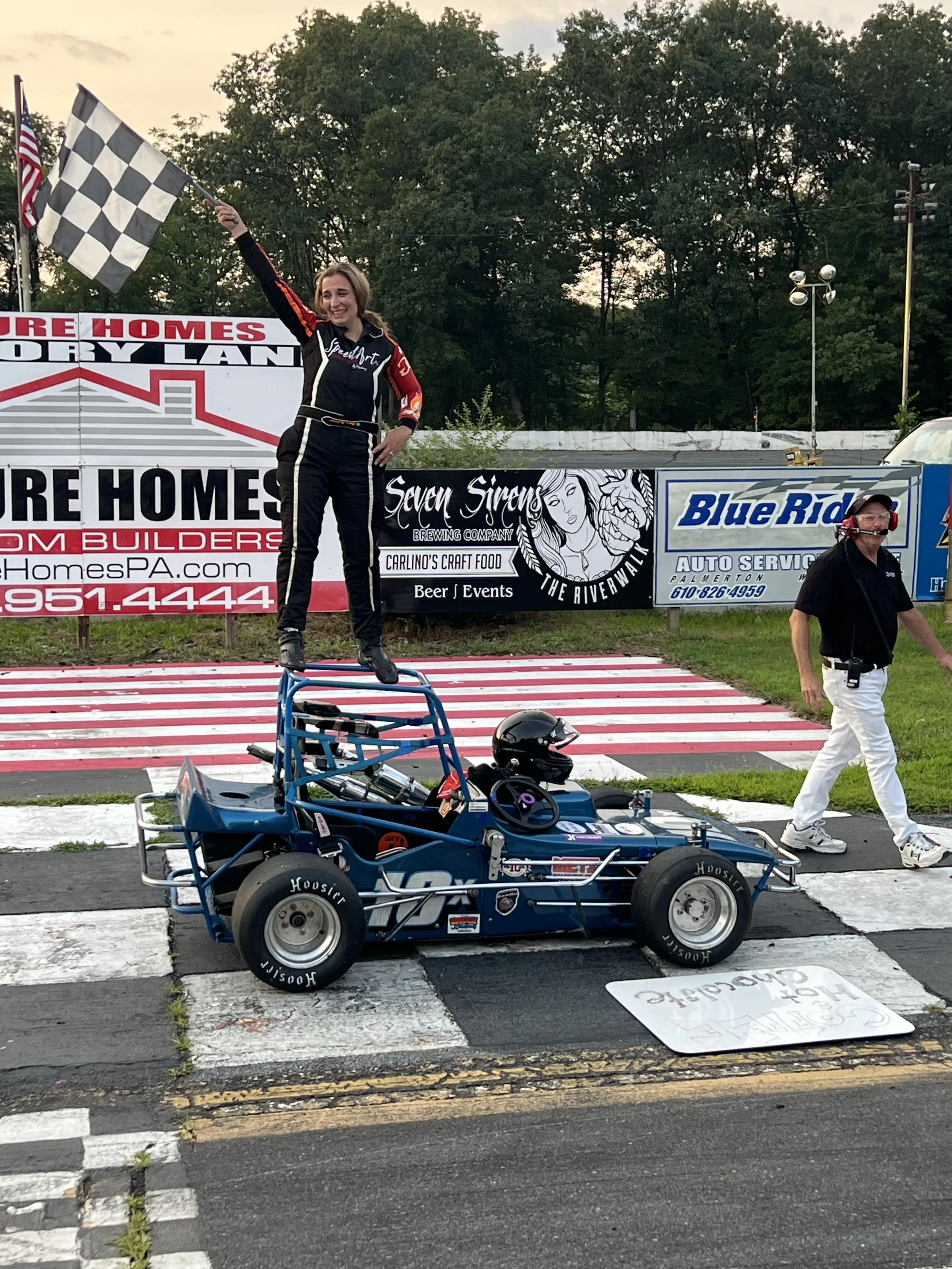 A woman in a racing suit standing on a winners' podium holding a checkered flag at a racetrack. A race car with a helmet next to it is in the foreground. There are contest banners and advertising signs in the background.