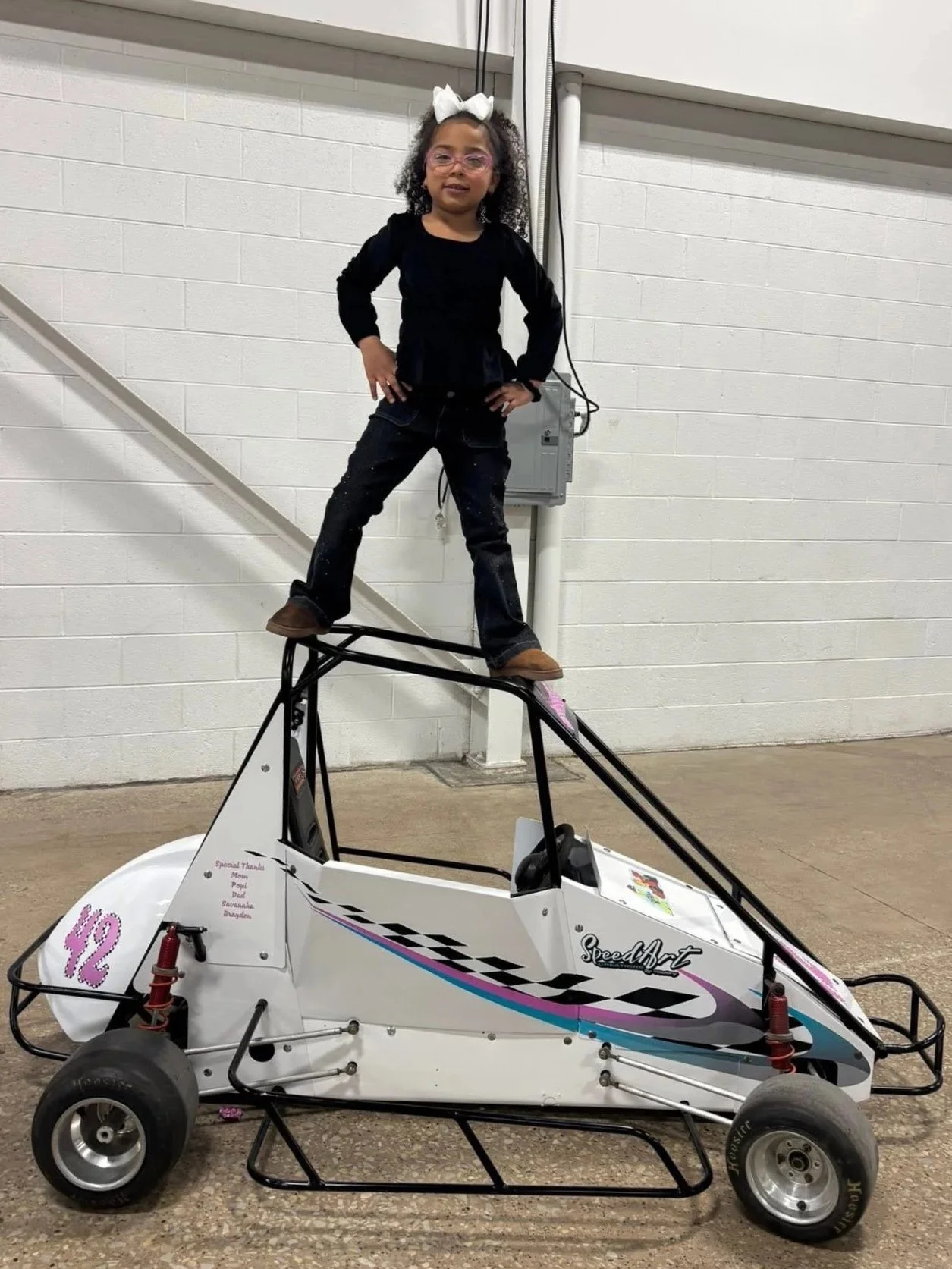 A young girl in glasses and a white bow on her head standing with her hands on her hips on top of a small race car inside a building with white brick walls.
