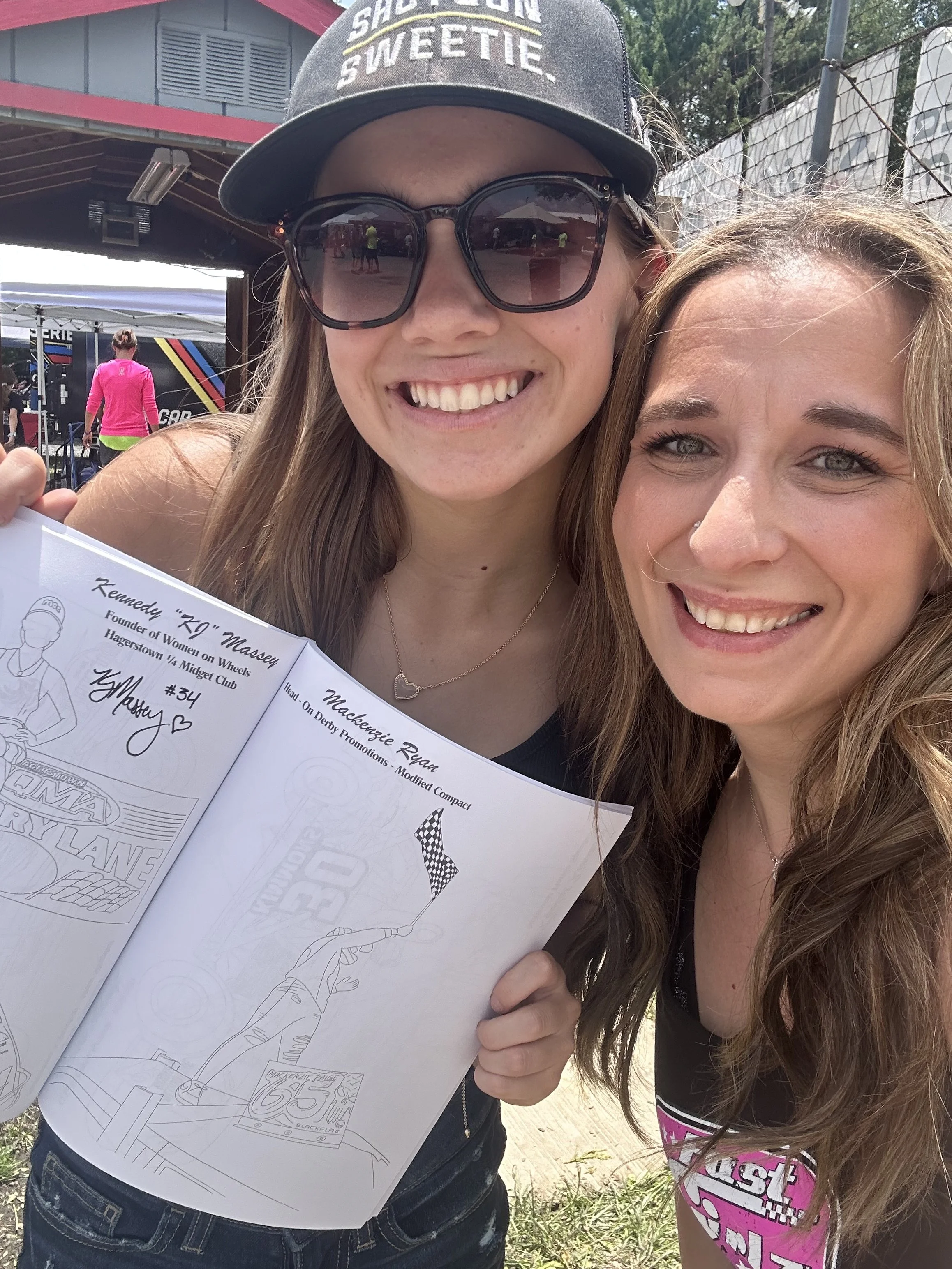 Two smiling women taking a selfie outdoors at a racing event. One woman is wearing sunglasses, a cap, and holding a coloring book. The other woman has wavy hair and is wearing a pink shirt with race-themed text. In the background, there are tents, a building, and a fence.