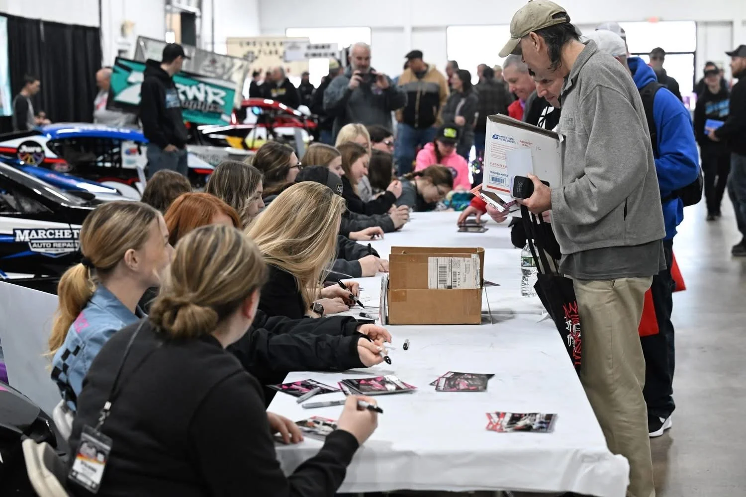 People gather at a table signing autographs, with race cars in the background at an event or convention.