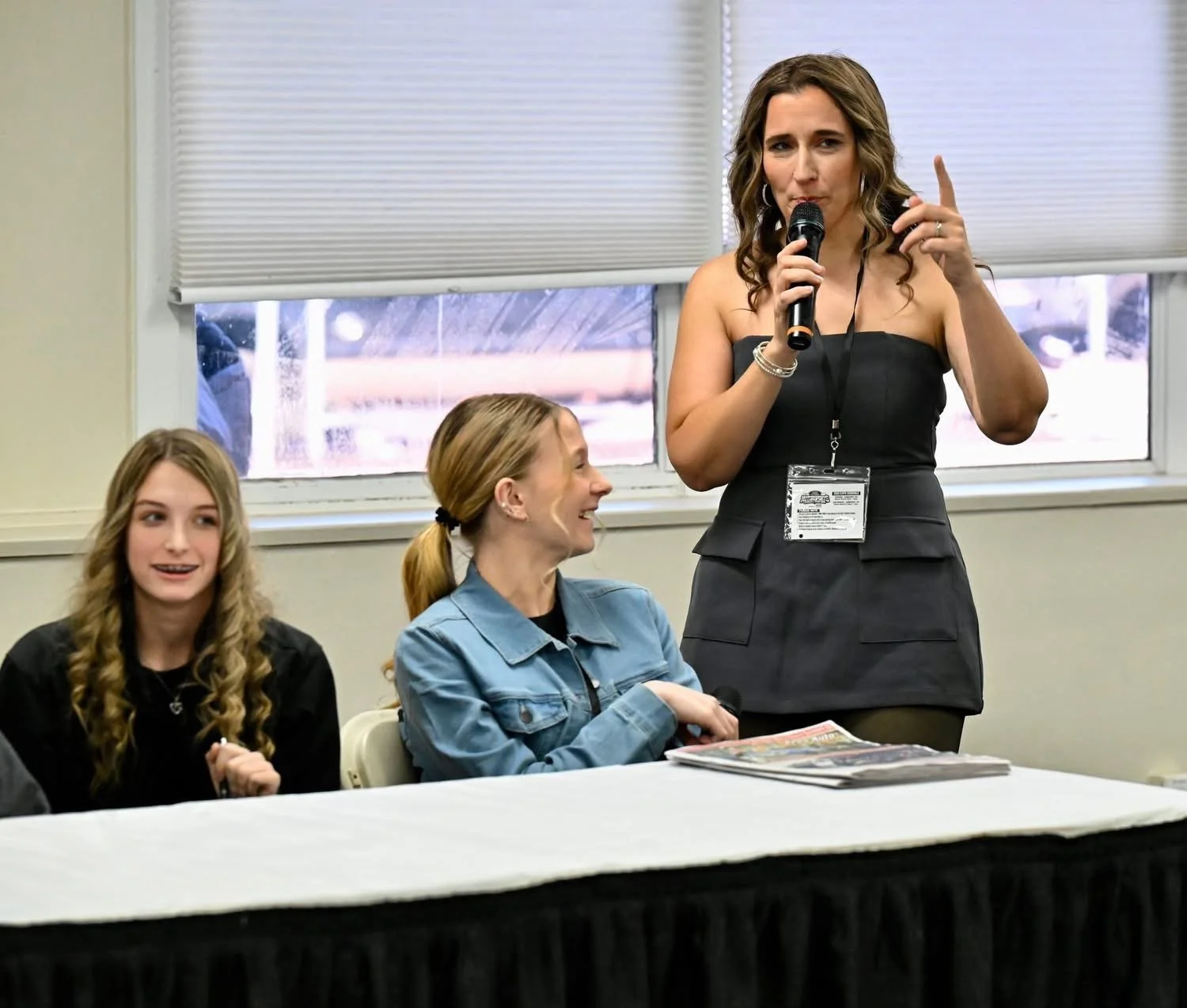 Three women sitting at a table during a panel or conference. One woman is standing, holding a microphone, and speaking or making a point. The woman in the middle is smiling and looking up at her, while the woman on the left is also smiling and looking forward. There are papers and magazines on the table.