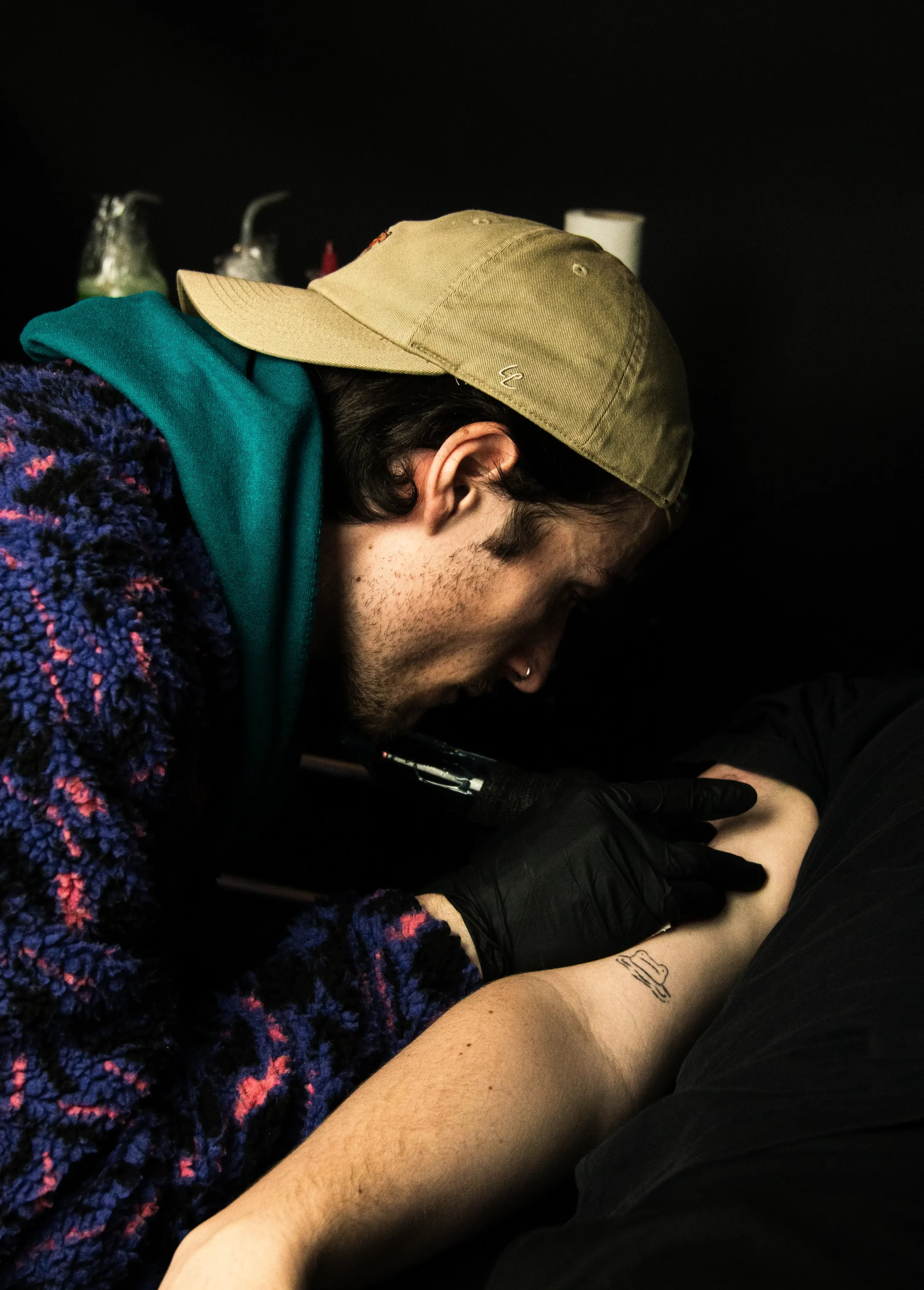 Eine Person mit dunklen Haaren und einem Tattoo am Unterarm, die eine Tätowierung anfertigt, trägt schwarze Handschuhe und liegt auf einem Tisch. Sie trägt eine bunte Jacke mit lila, blau und pink Tönen, eine grüne Kapuzenjacke, eine beige Mütze und einen Ohrring in der linken Ohrmuschel.