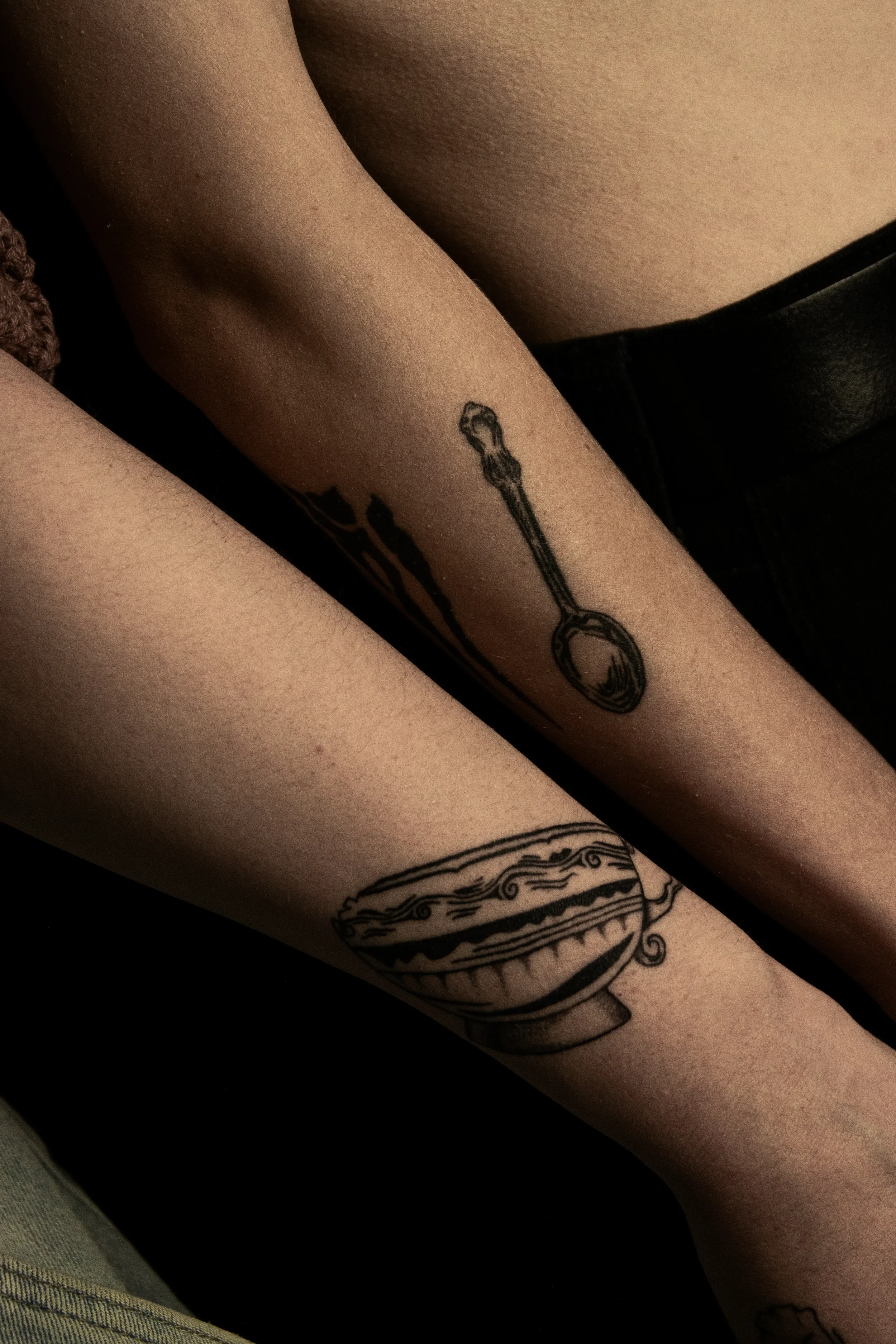 Nahaufnahme von Armen mit Tattoos, darunter eine Teekanne und ein Löffel im schwarzen Tattoo-Stil.