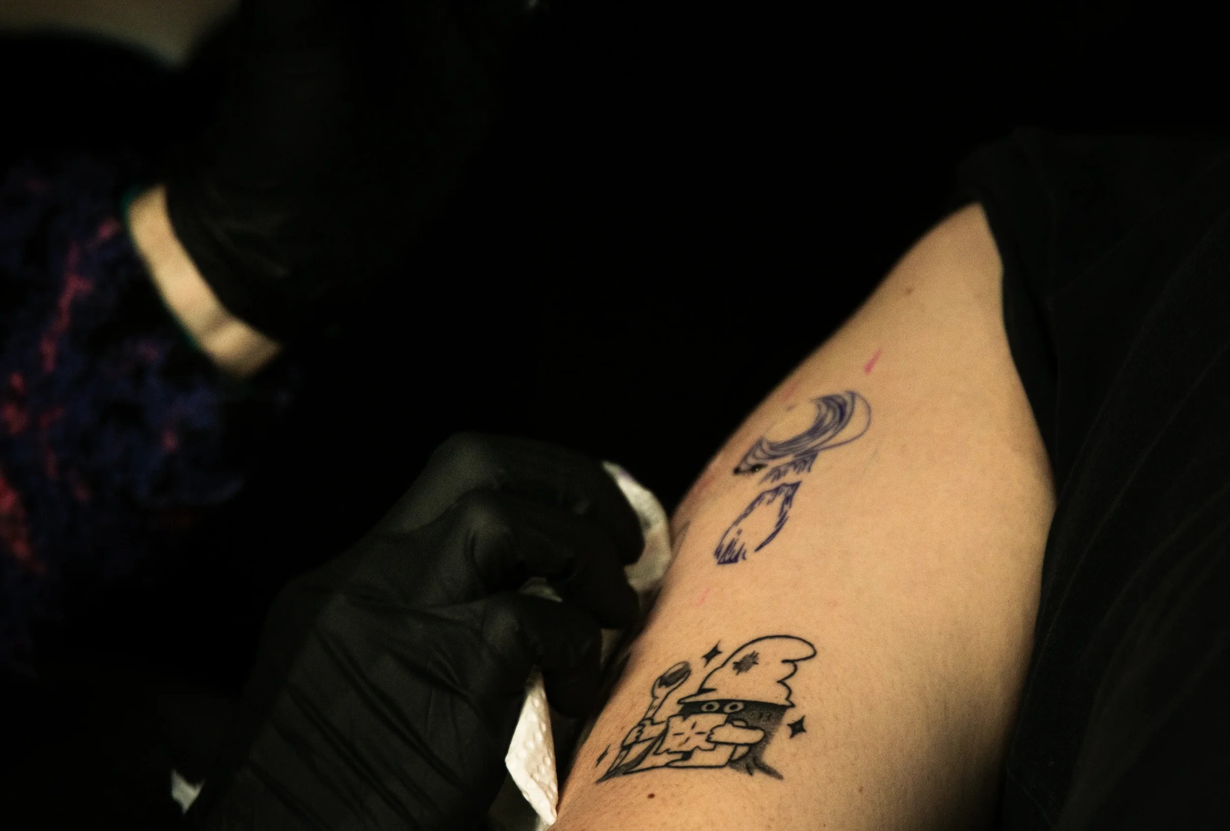 Nahaufnahme eines Unterarms mit mehreren Tattoos, während eine Tätowiererhand ein Tattoo absolviert.