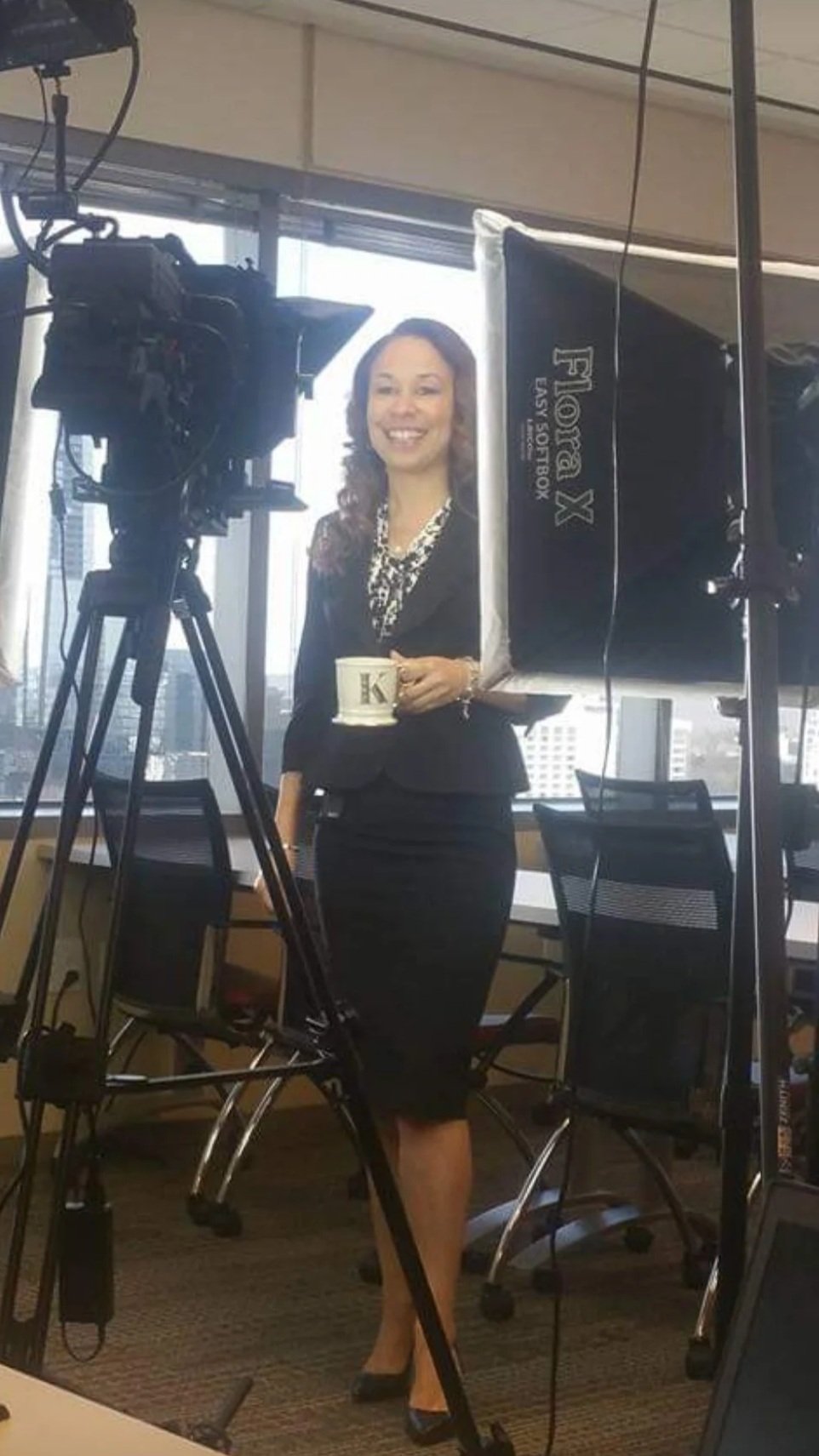Kelley as a spokesperson, holding a coffee mug, standing in front of a camera and studio lighting in a modern office with large windows and cityscape view.