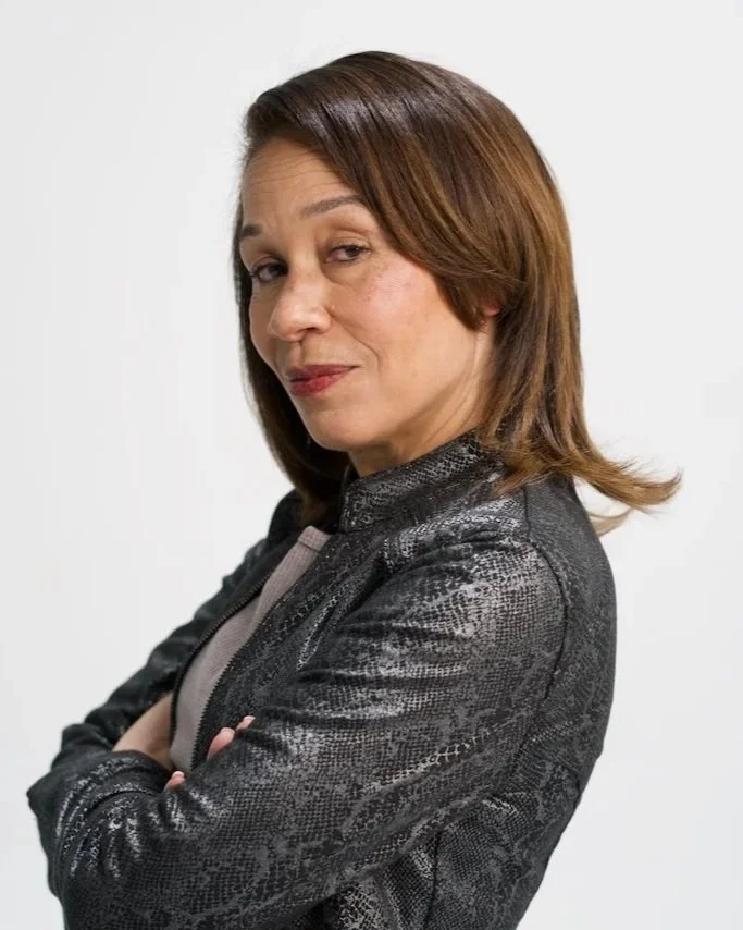 Kelley Powell on camera look with shoulder-length brown hair, wearing a black and gray snakeskin-patterned jacket, with her arms crossed and looking at the camera.
