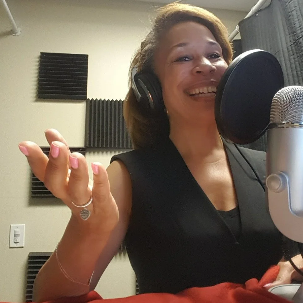 Kelley Powell in a recording studio, wearing headphones, smiling, seated near a microphone with soundproofing foam on the wall behind her.