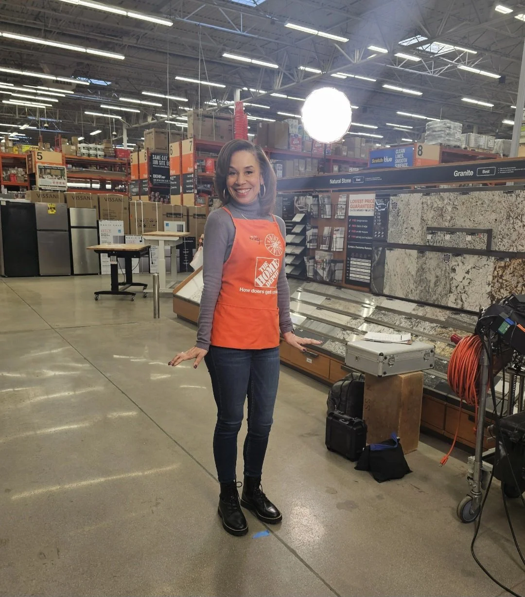 Kelley standing in a hardware store, smiling, wearing a grey top, jeans, black boots, and an orange apron with the Home Depot logo, surrounded by shelves of building materials and appliances.