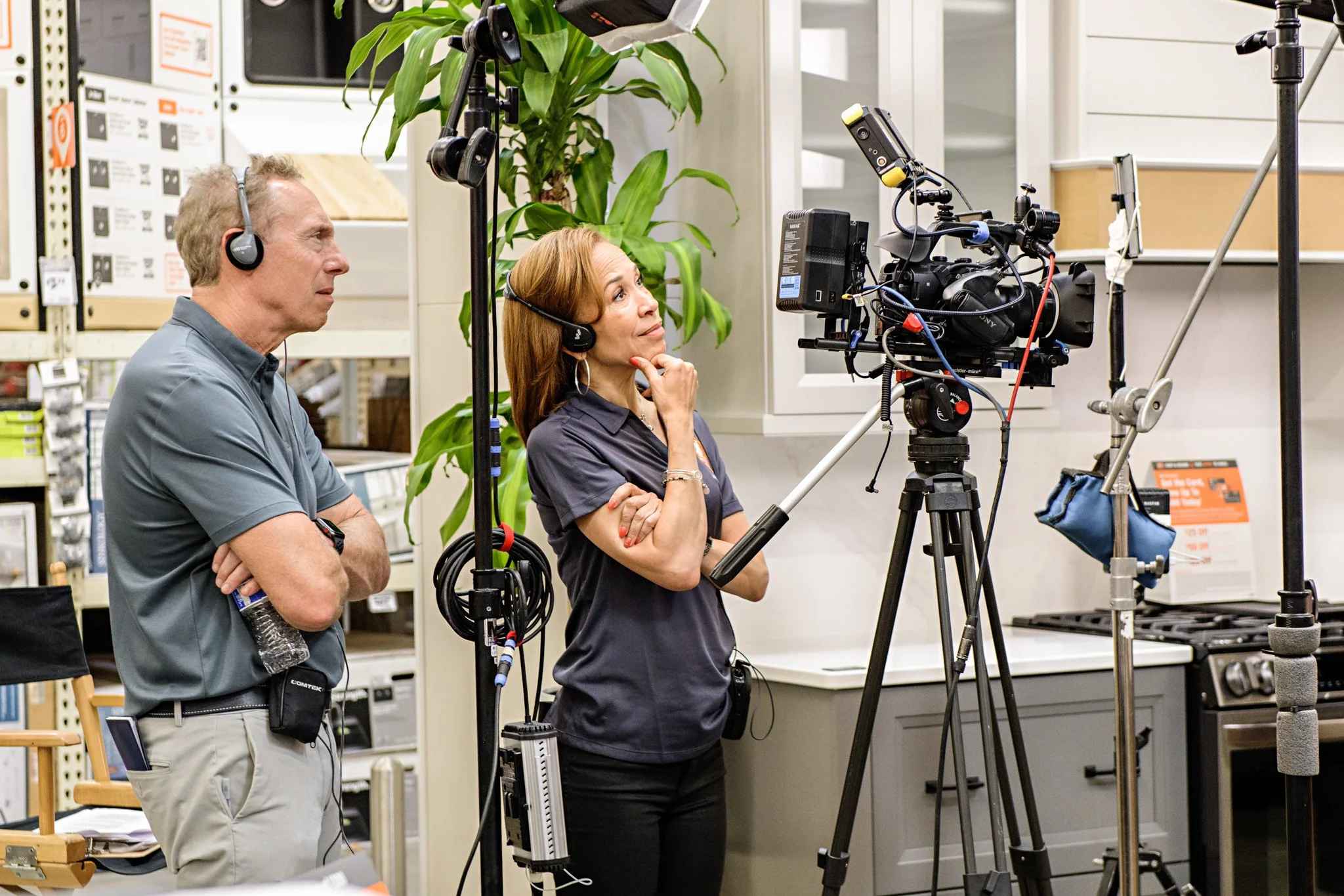 Kelley and Brian are working with film equipment in a storage or warehouse setting. They are both wearing headsets and looking at a camera mounted on a tripod, with shelves and boxes visible in the background.