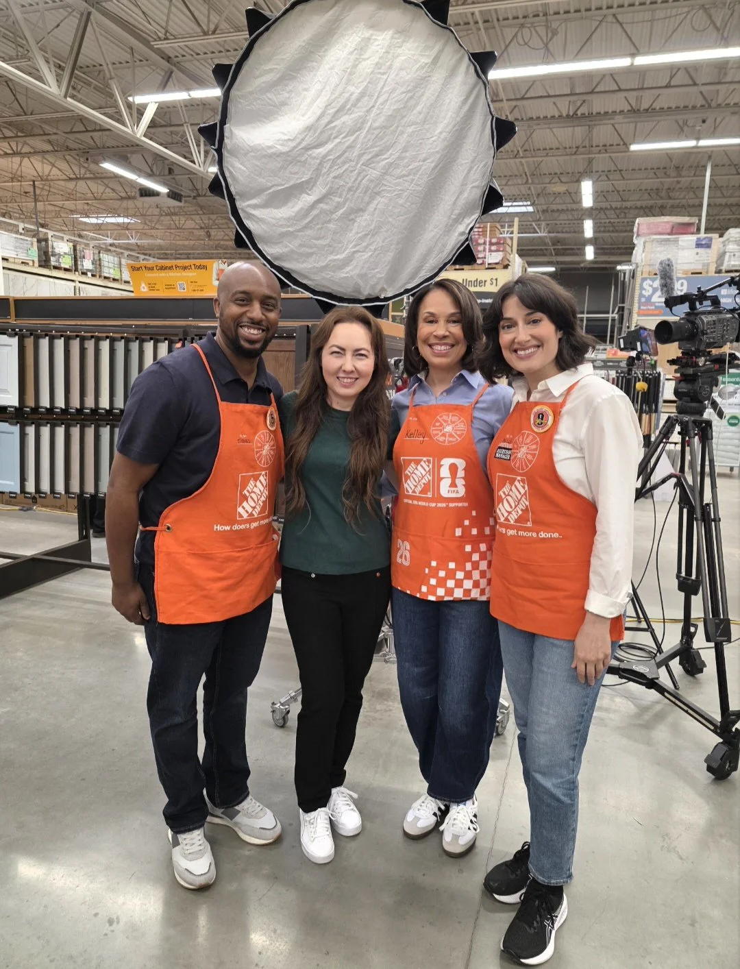 Kelley inside a hardware store with displays of flooring and tools in the background. One woman and one man are wearing orange aprons with the Home Depot logo. A large professional light is above them, and there is a camera on a tripod to the right.