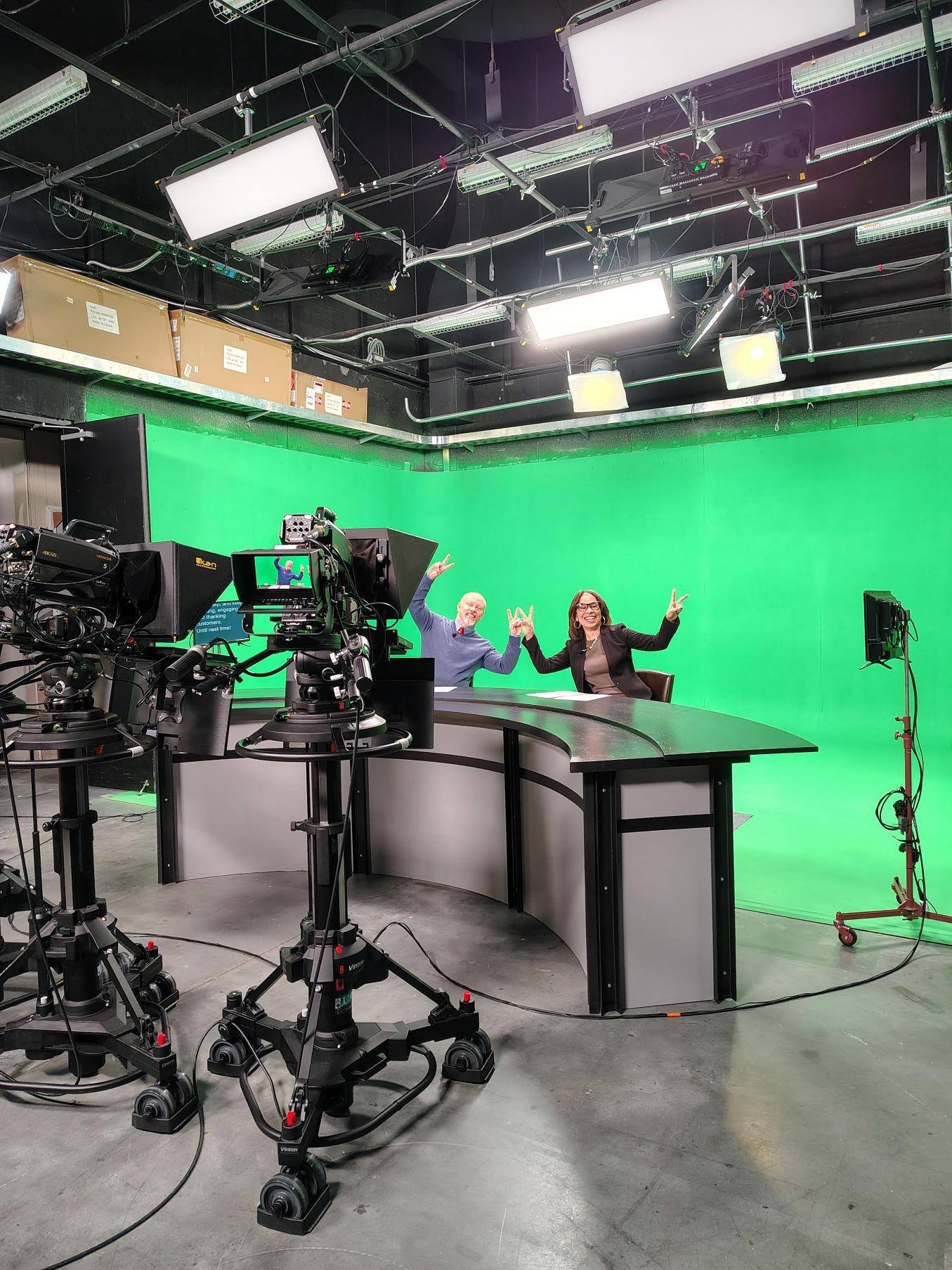 Kelley sitting at a news desk in a television studio with a green screen background. They are smiling and making peace sign gestures. There are multiple cameras and studio lights overhead.