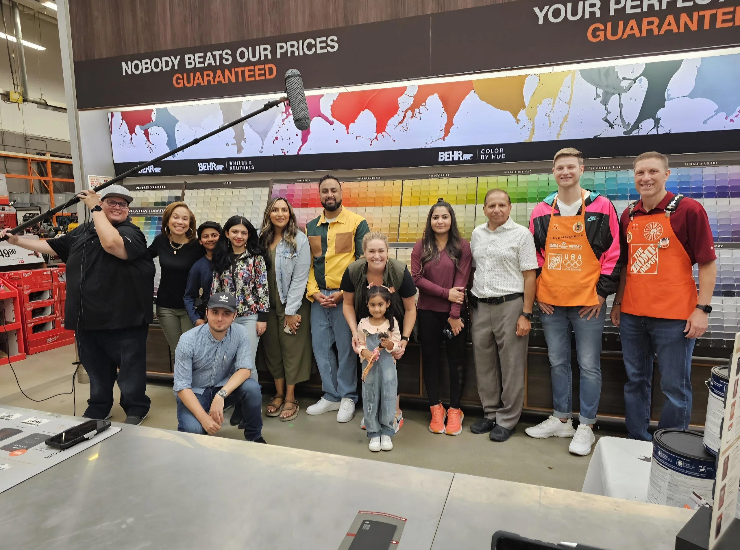 Group of people standing and kneeling in front of paint color displays inside a store, with a large sign above reading 'Nobody beats our prices guaranteed' and 'Your perfect guarantee'.