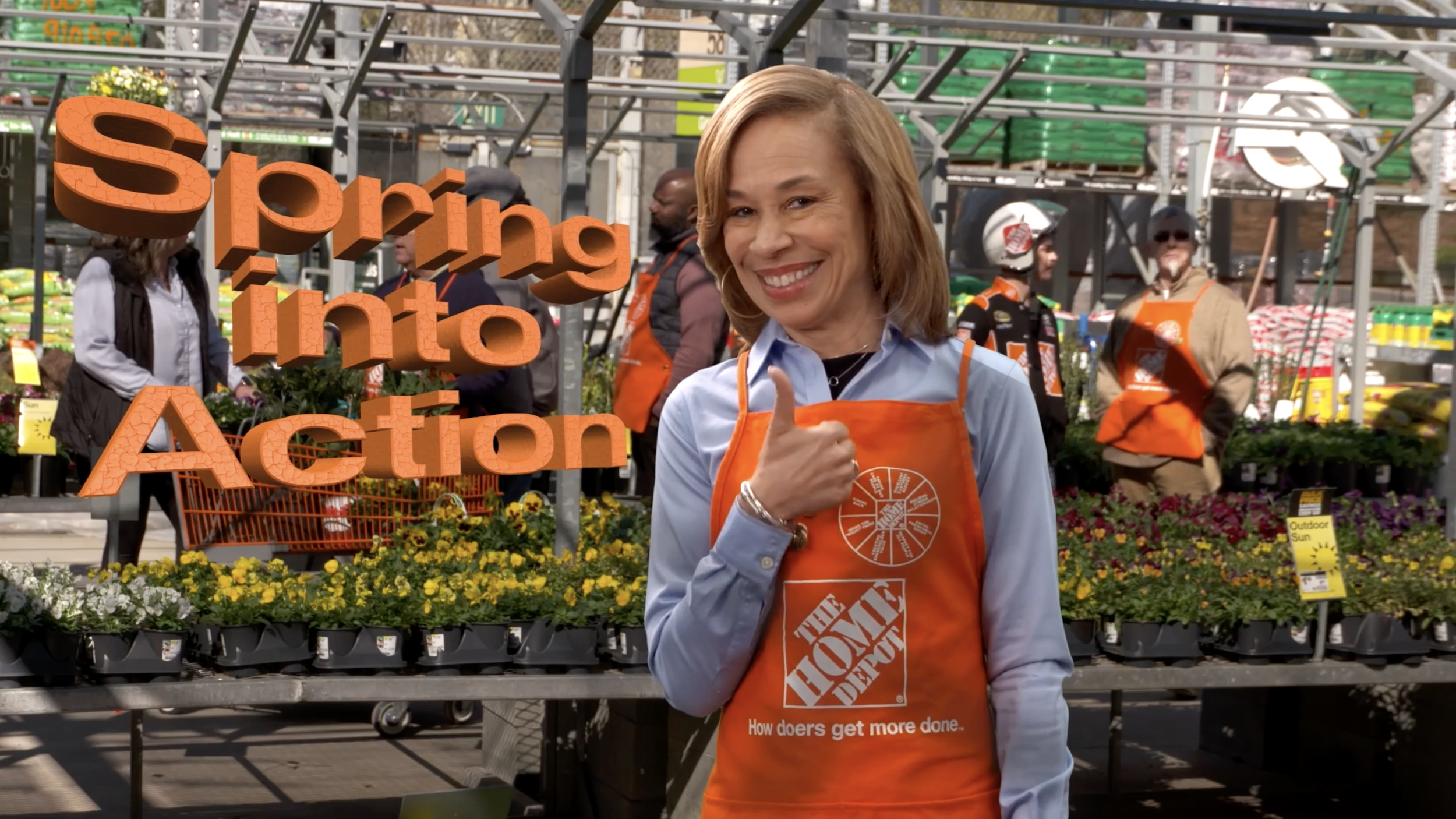 Kelley in an orange Home Depot apron giving a thumbs up at a garden section of a home improvement store, with colorful flowers and garden supplies in the background.