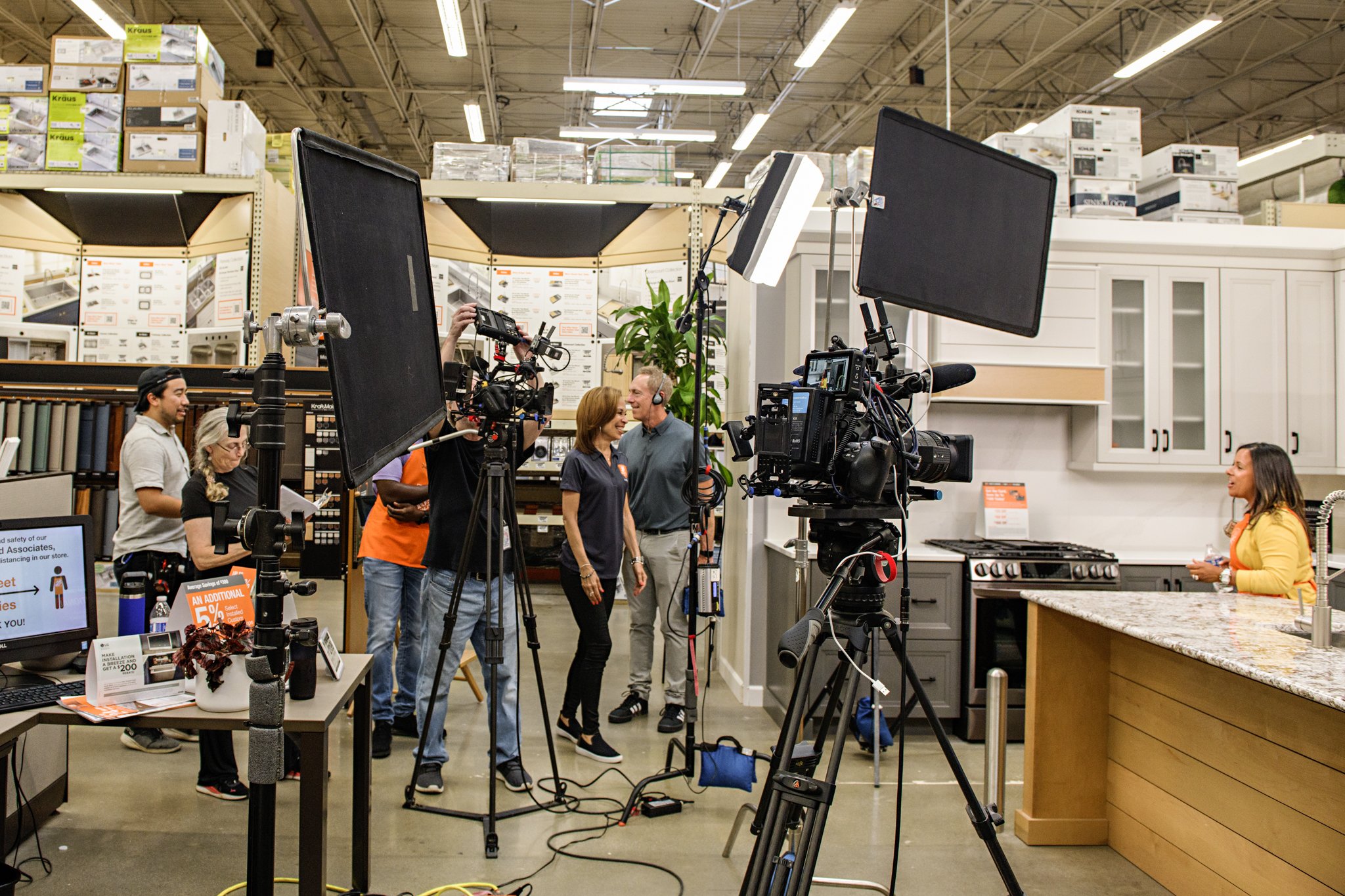 Filming setup inside a home improvement store with multiple camera and lighting equipment, several people including a woman being interviewed or photographed, and store shelves with kitchen and home products in the background.