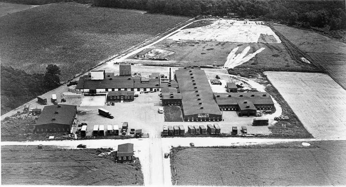 Aerial view of a large industrial building complex with multiple structures, parking lots, and surrounding farmland.