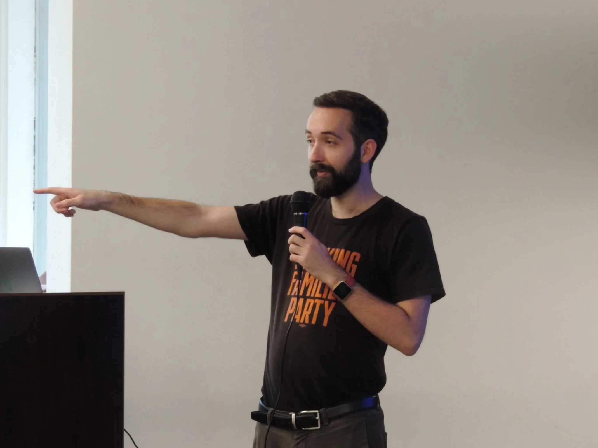 A man with dark hair and beard, wearing a black T-shirt with orange text, is holding a microphone in his right hand and pointing with his left hand towards a white wall. The room has a window on the left side letting in natural light.