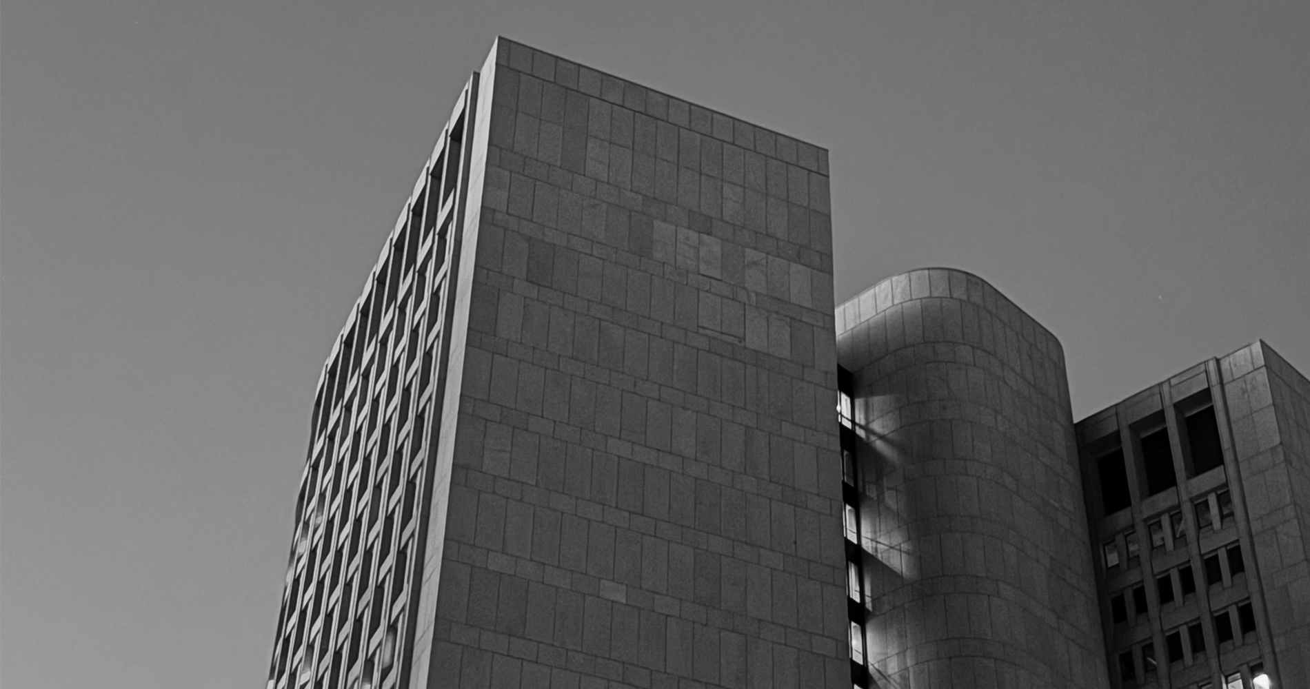 A black and white photograph of a modern high-rise building with a unique architectural design, featuring rectangular and curved sections, and windows running vertically along one side.
