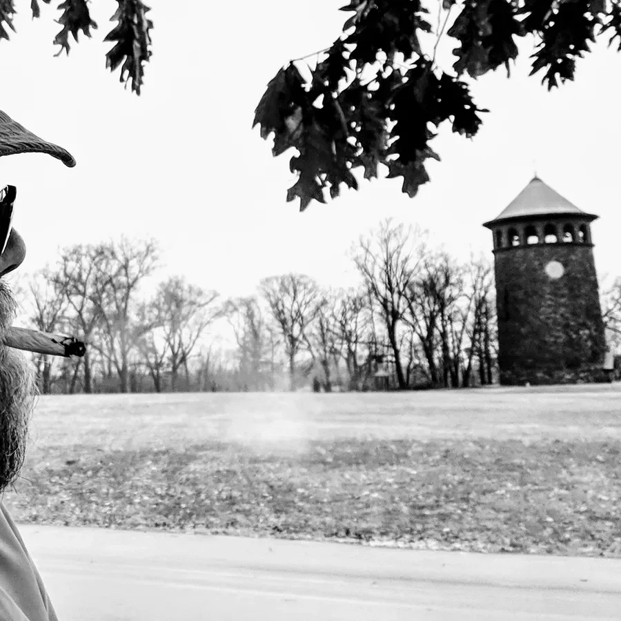 Black and white photo of a man smoking a cigar, with trees and a tower in the background.