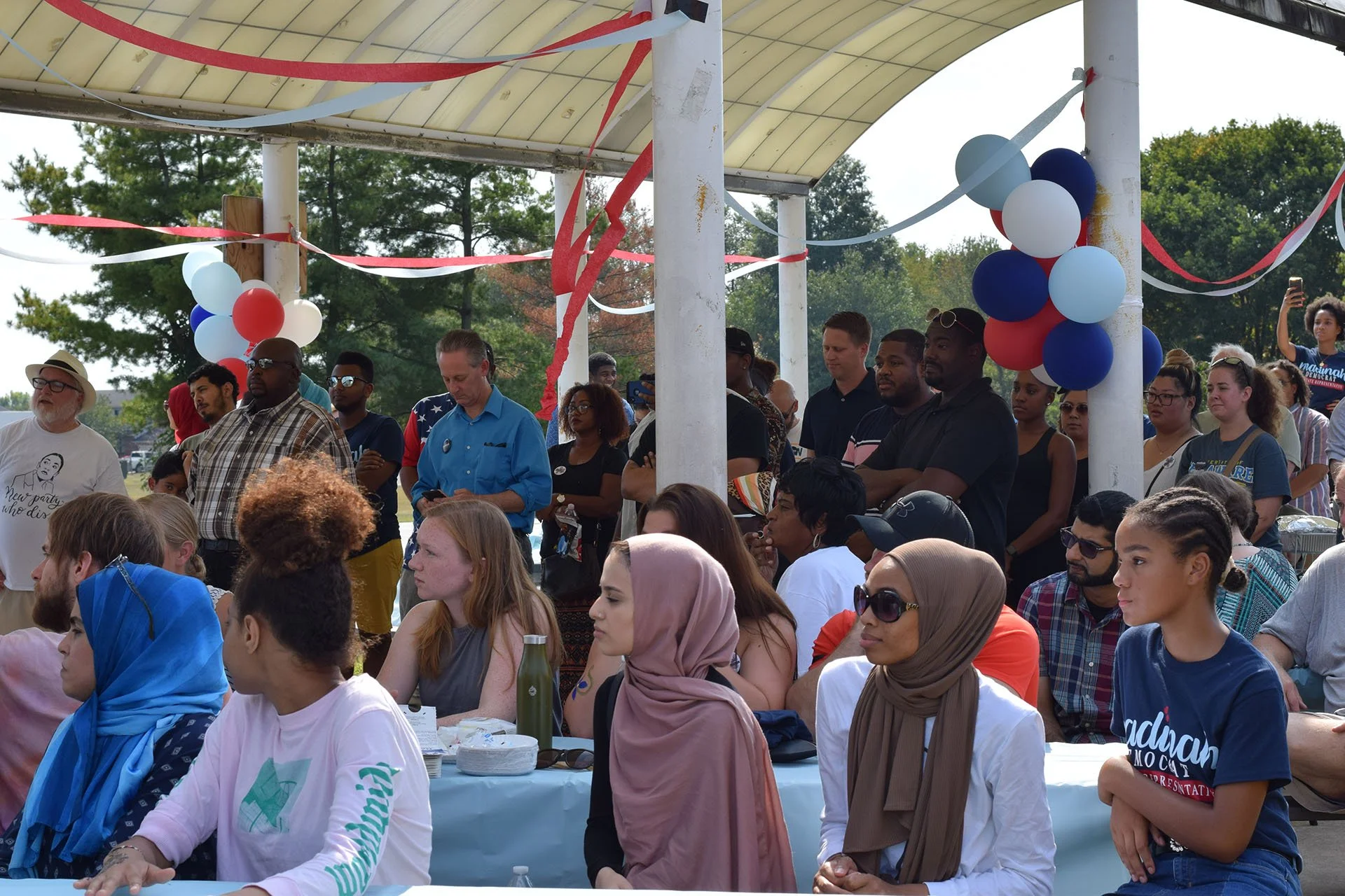 Crowd gathered at an outdoor event under a canopy decorated with red, white, and blue balloons and streamers. Some individuals are sitting at a table, while others stand listening, with diverse ages and ethnicities.