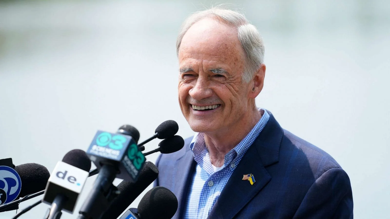 An older man in a blue suit and light blue shirt smiling at a microphone at a press conference outdoors.