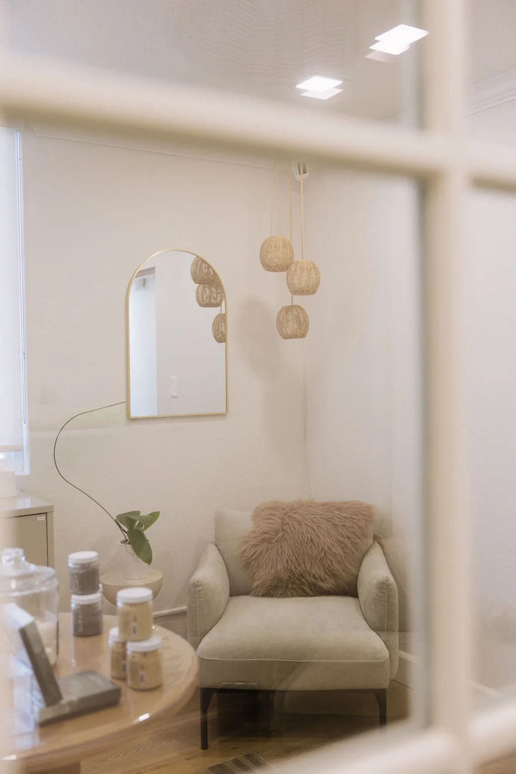 A cozy, minimalist living room corner with a light green armchair, a fluffy pink pillow, a small side table with jars, a modern gold-framed mirror, and hanging woven pendant lights, all in neutral tones.