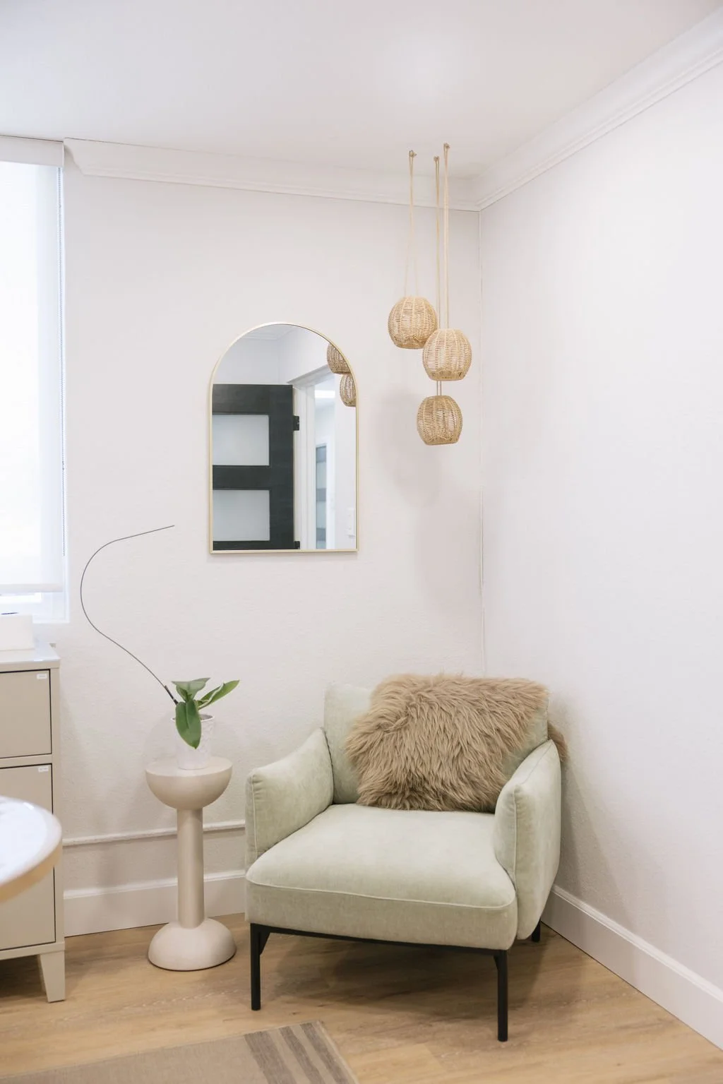 A cozy corner with a plush beige armchair with black metal legs, a tan fluffy pillow, and a small white side table with a green plant. A mirror and hanging wicker lamps are on white walls, and a window with white blinds is on the left.