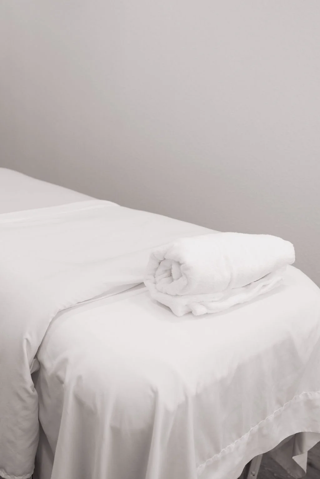 A neatly made bed with white sheets and a pillow, topped with a rolled white towel, against a plain off-white wall.