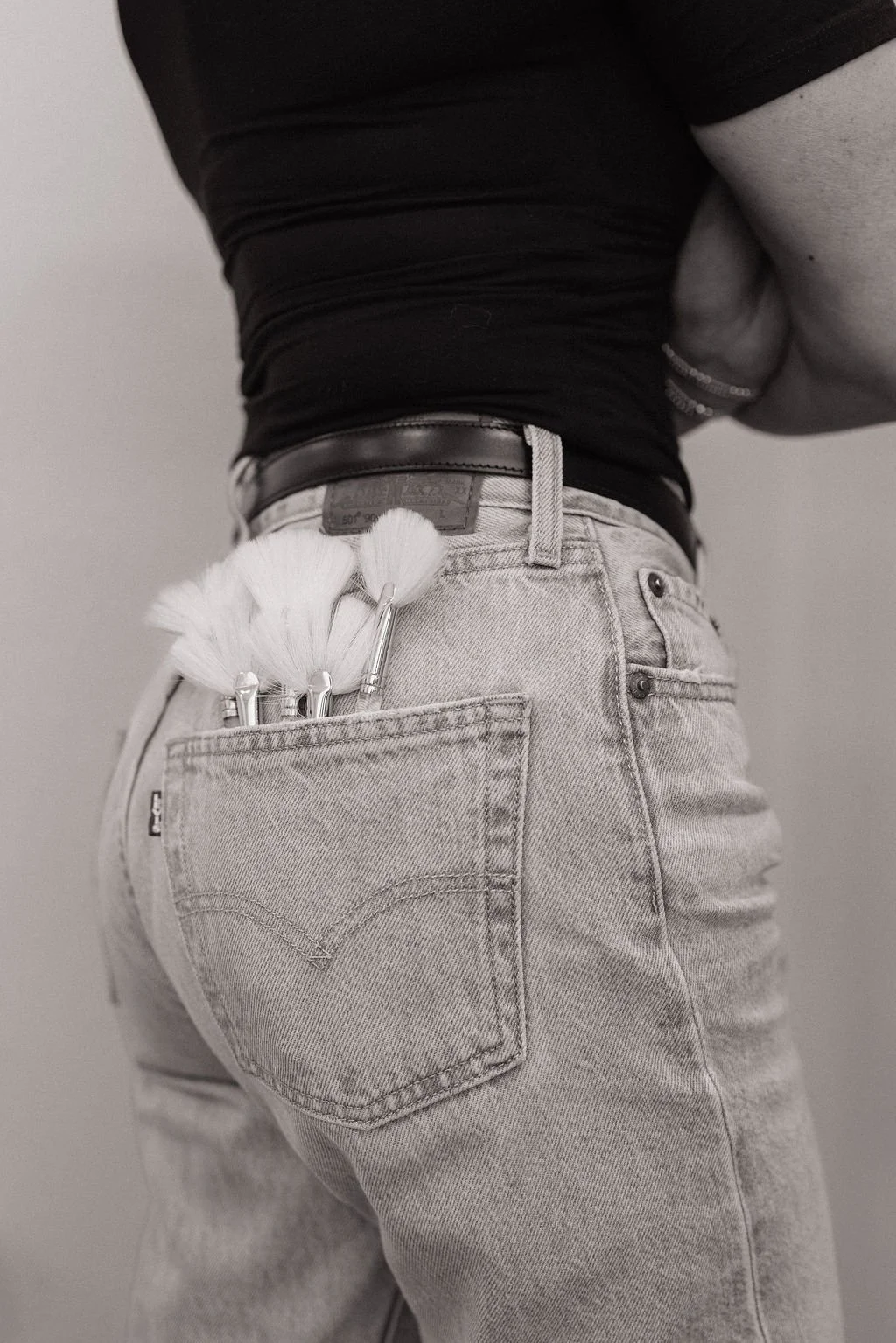 Person wearing light jeans with a pocket holding paintbrushes, black belt, black shirt, and gray skin, standing against a plain wall.