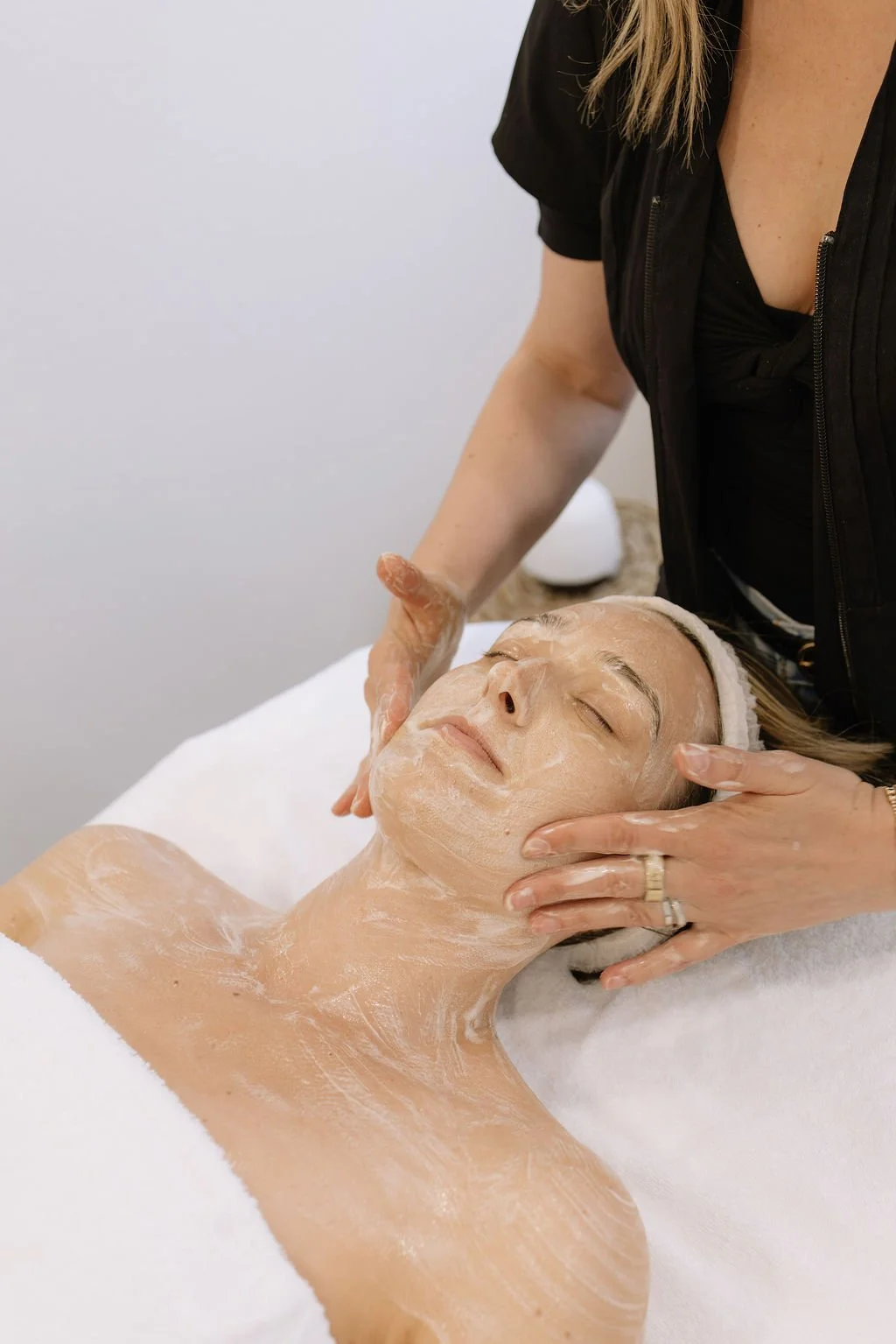 A woman receives a facial treatment from a skincare specialist in a spa setting, lying on a treatment table with her eyes closed.