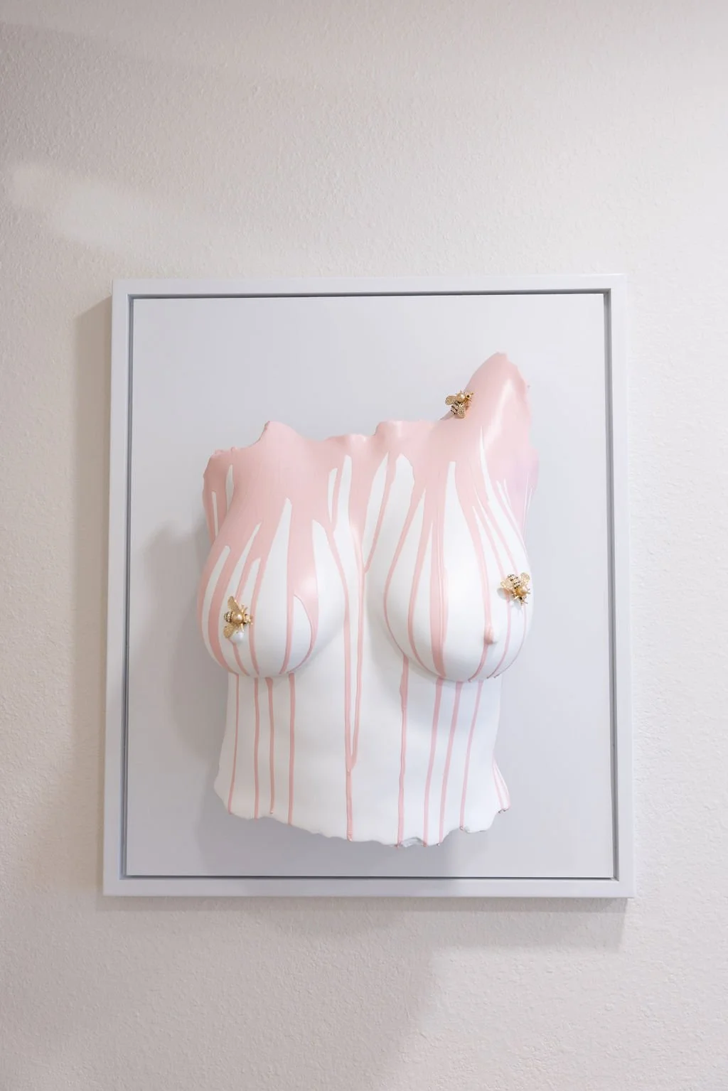 Mixed media artwork of a stylized female torso with pink and white striped paint drips, adorned with three small gold bee sculptures.