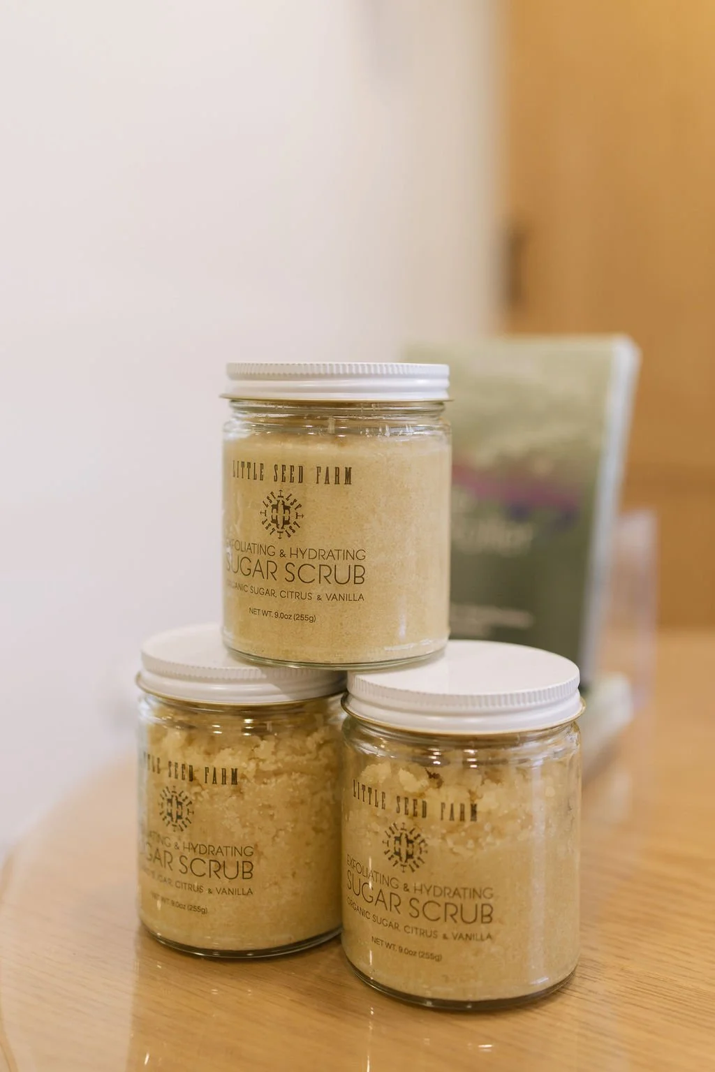 Three jars of exfoliating and hydrating sugar scrub with organic sugar, citrus, and vanilla, placed on a wooden surface, with a blurred green bag in the background.