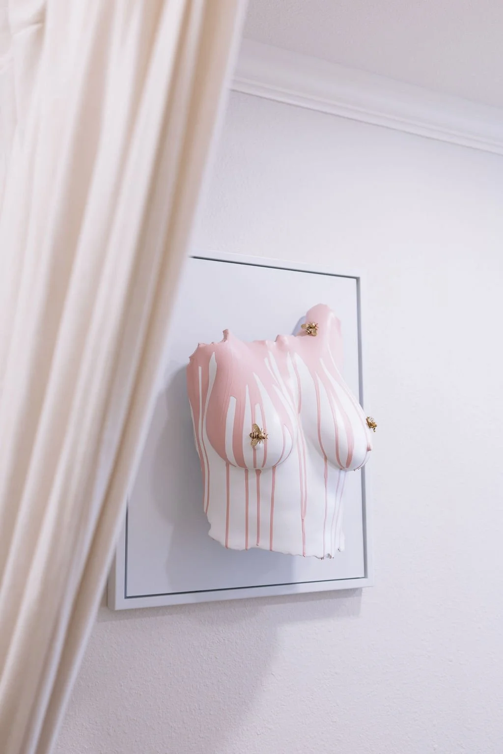 An art piece featuring a wall sculpture of a female torso with pink and white drip paint, adorned with small metallic bees, mounted on a white wall with crown molding.