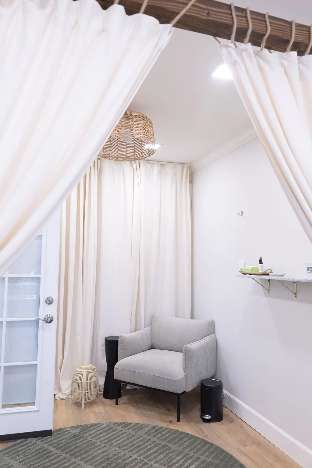 A cozy interior corner with a light gray armchair, black side tables, white curtains, and a wall-mounted shelf with toiletries. A jade rug covers part of the wooden floor, and a wicker lampshade hangs from the ceiling.