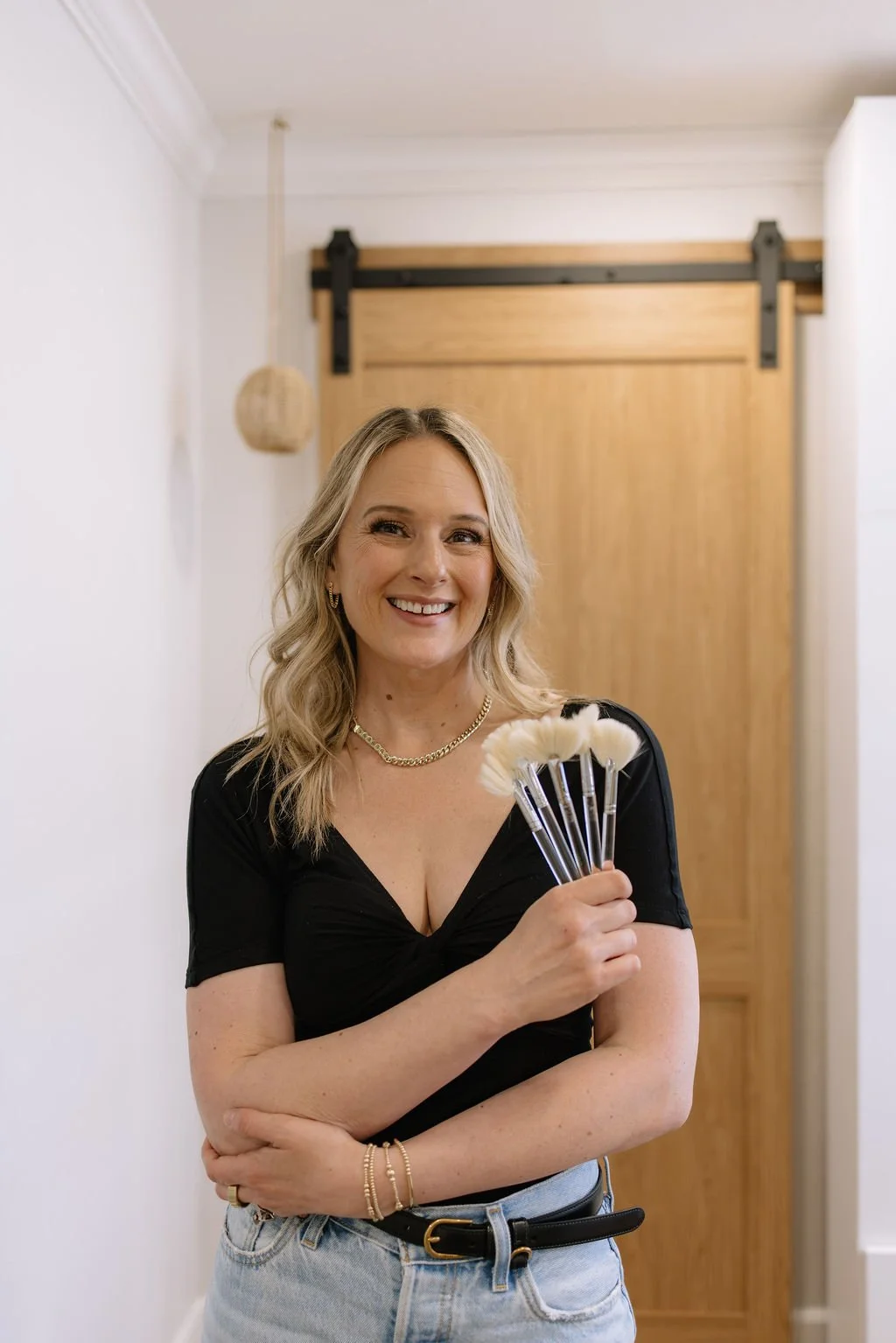 A smiling woman with blonde hair holding a handful of makeup brushes, standing in a room with a wooden sliding door in the background.