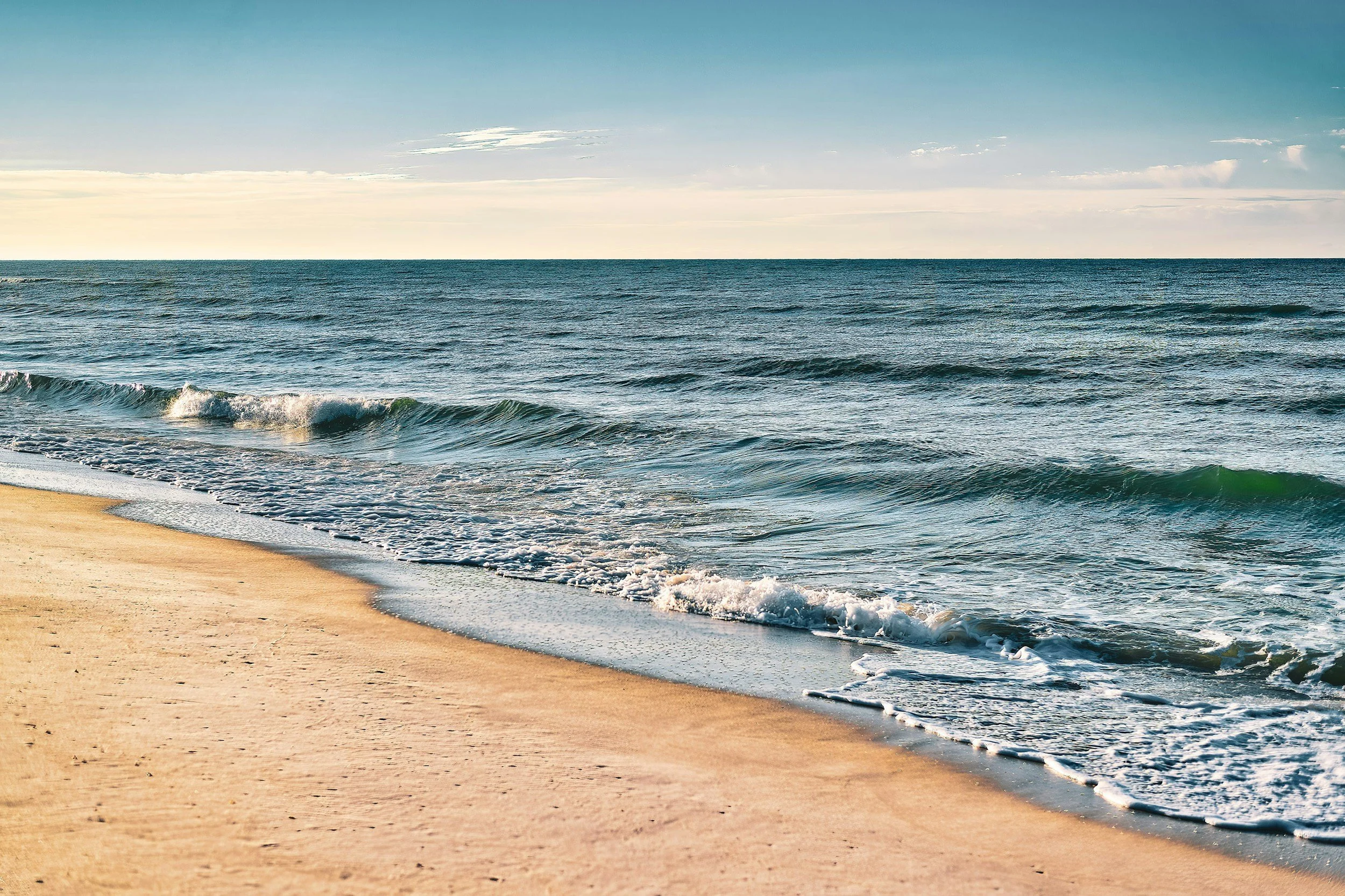 A serene view of the ocean with gentle waves hitting a sandy beach under a clear sky.