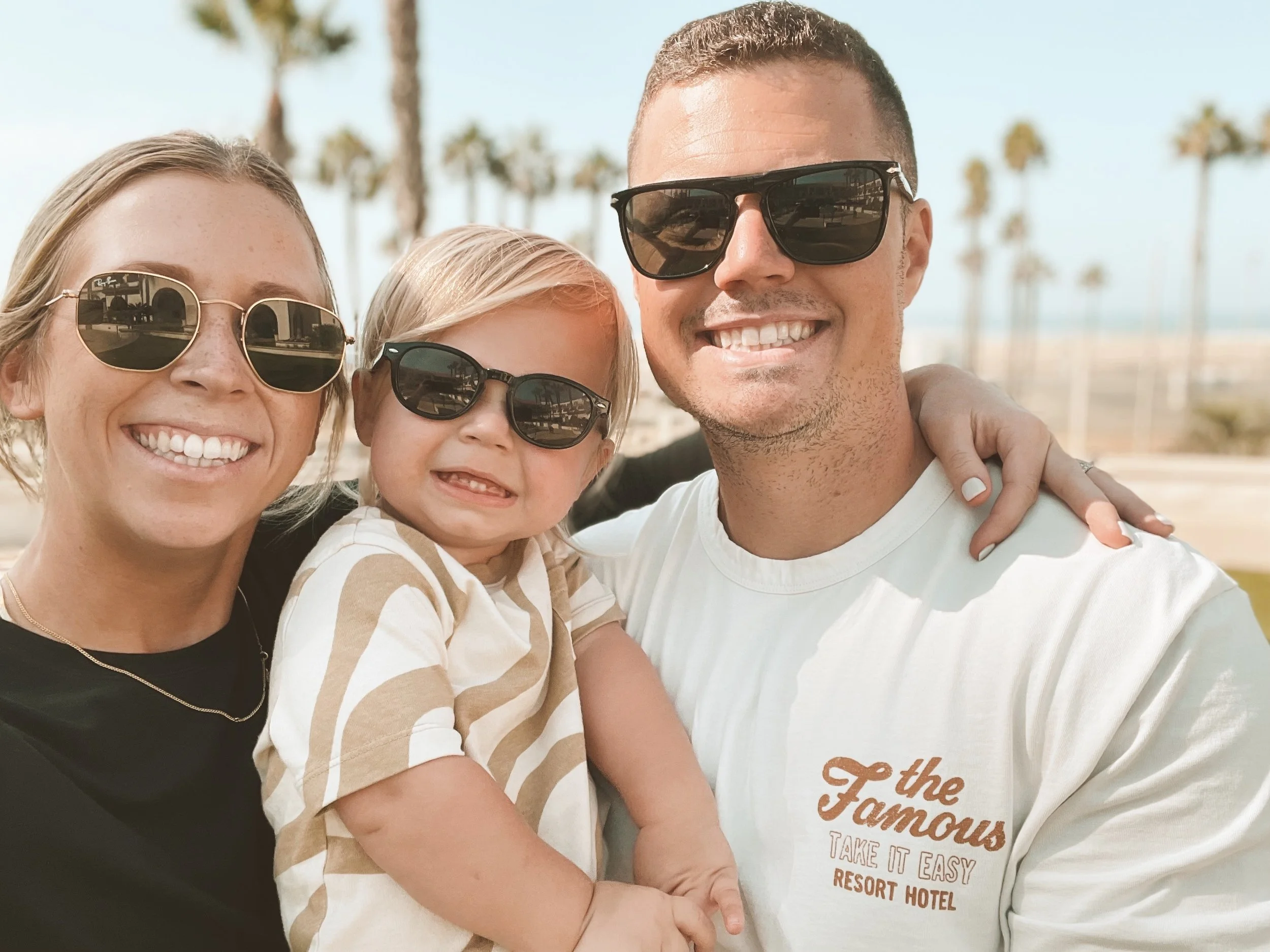 A group of three people smiling outdoors near palm trees, with one women and one man and one child all wearing sunglasses and close together, appearing happy.