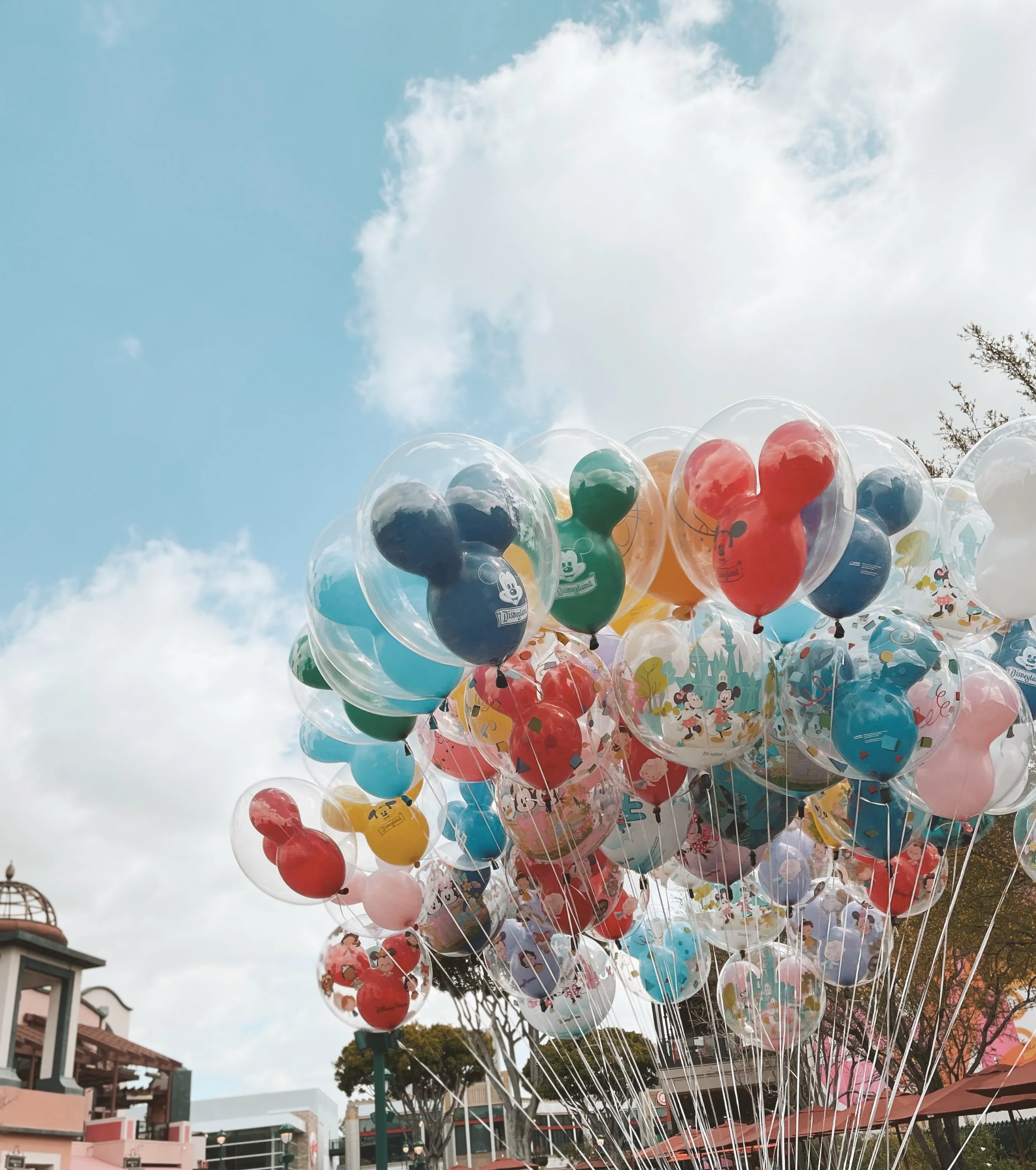 Colorful balloons featuring Disney characters, floating in the sky above an outdoor area with buildings and trees, possibly at an amusement park or festival.