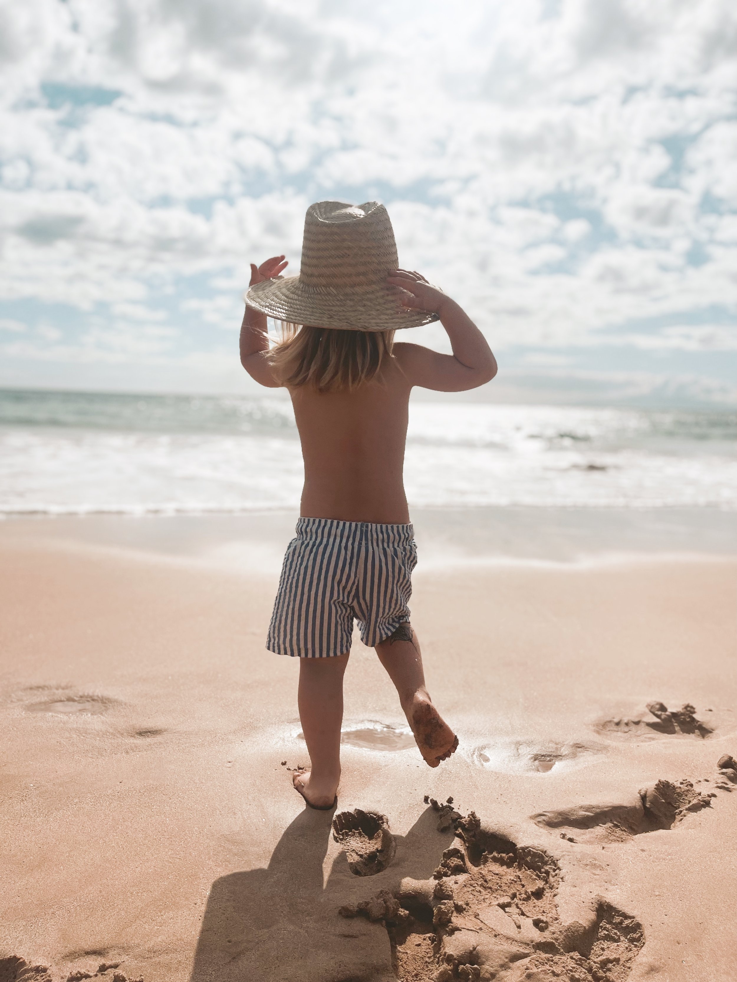 A child walking on the beach holding a wide-brimmed straw hat, wearing striped shorts, as footprints are visible in the sand. The ocean and cloudy sky are in the background.