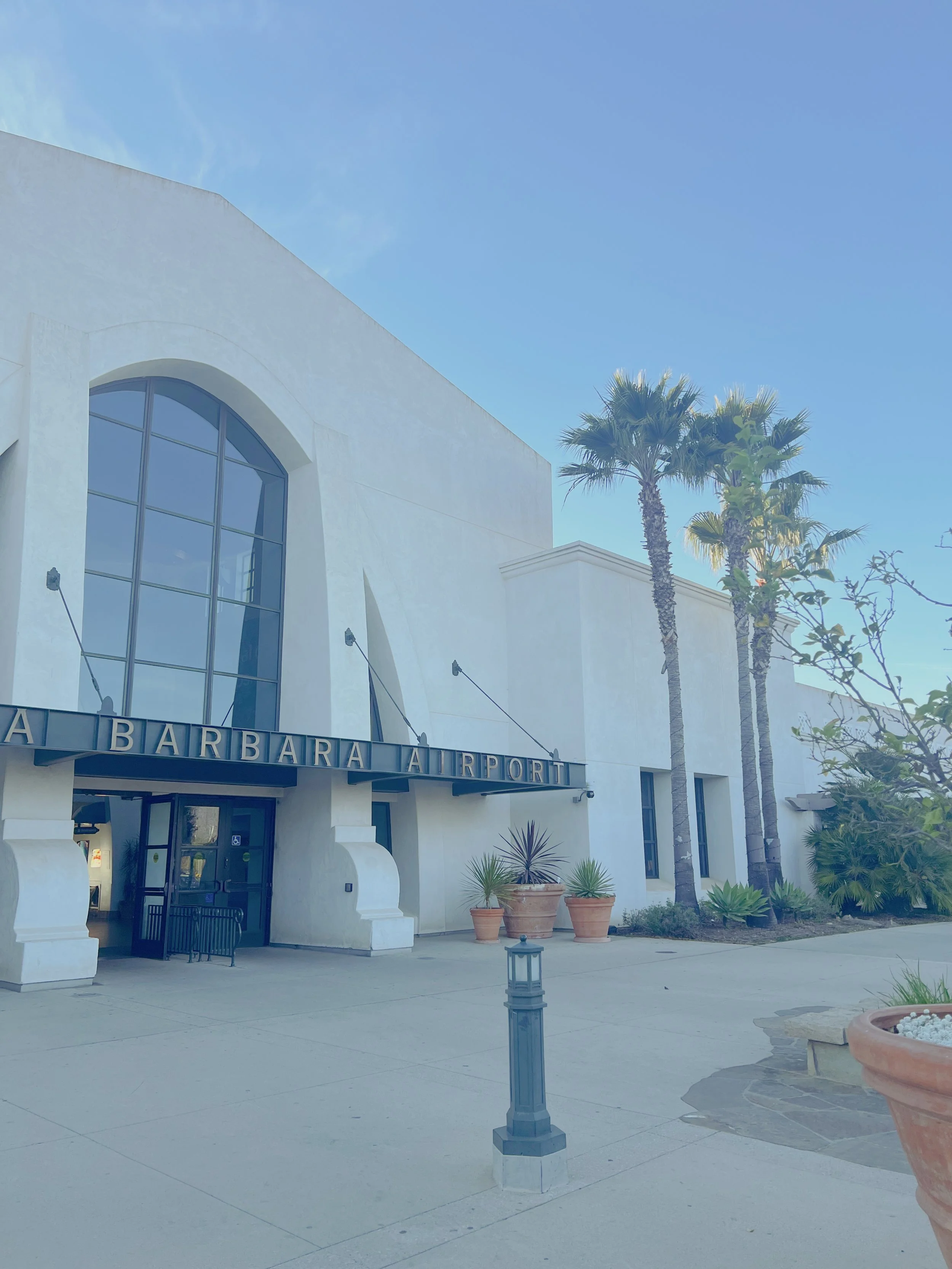 Facade of a white building with arched windows and the sign 'SANTA BARBARA AIRPORT'. There are tall palm trees, potted plants, and a lamp post in the foreground on a clear, sunny day.