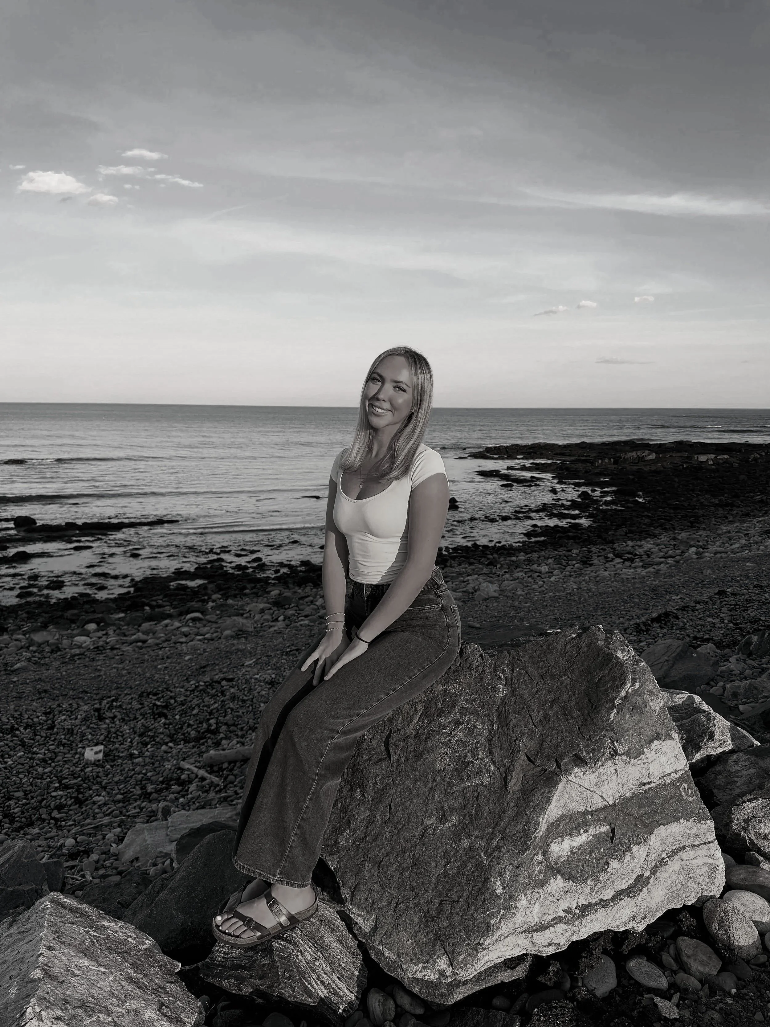 A woman sitting on a large rock at a rocky shoreline, smiling at the camera, with the ocean and sky in the background, in black and white.