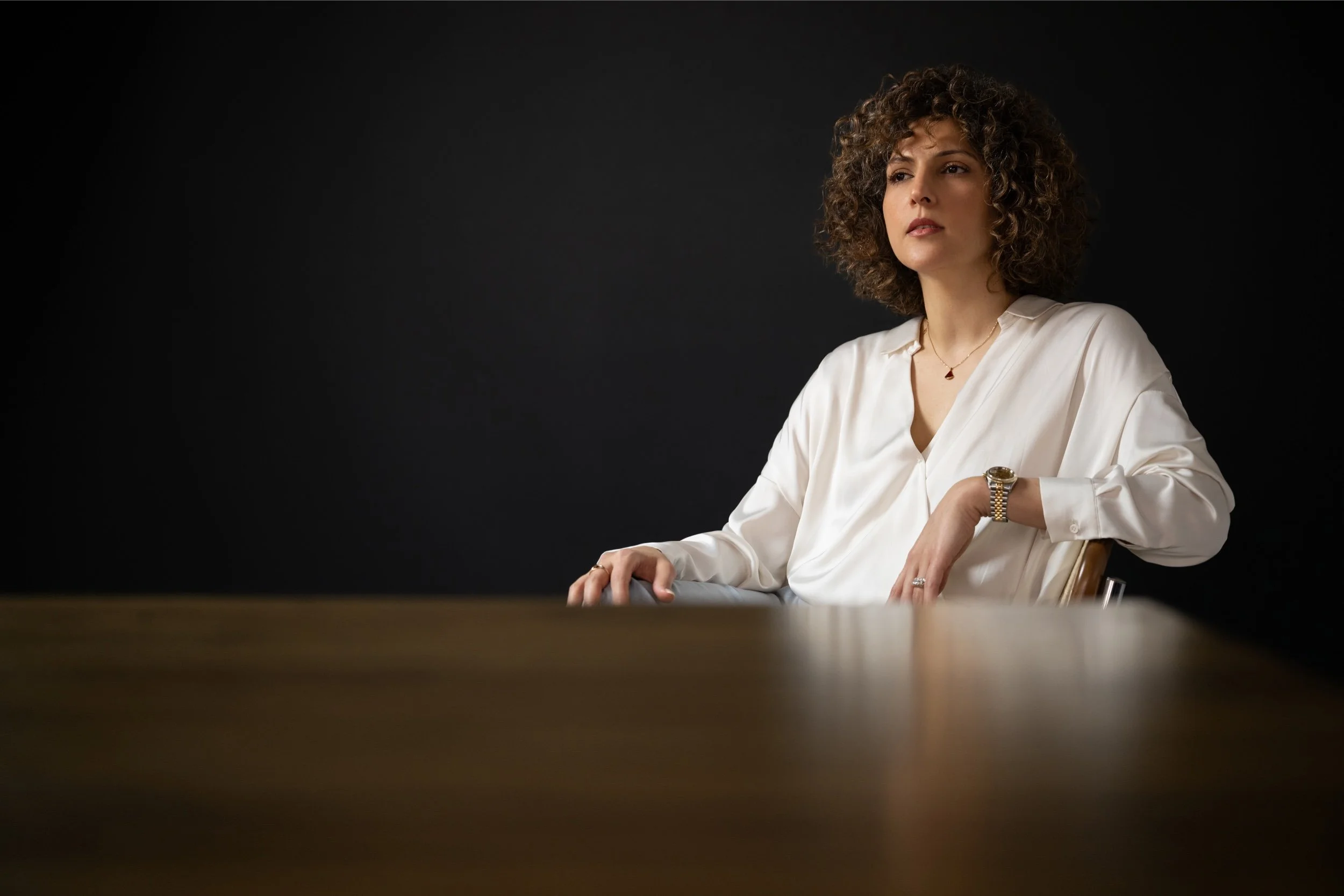 A woman with curly hair sitting at a table, wearing a white blouse, gold watch, ring, and a necklace with a small pendant, against a dark background.